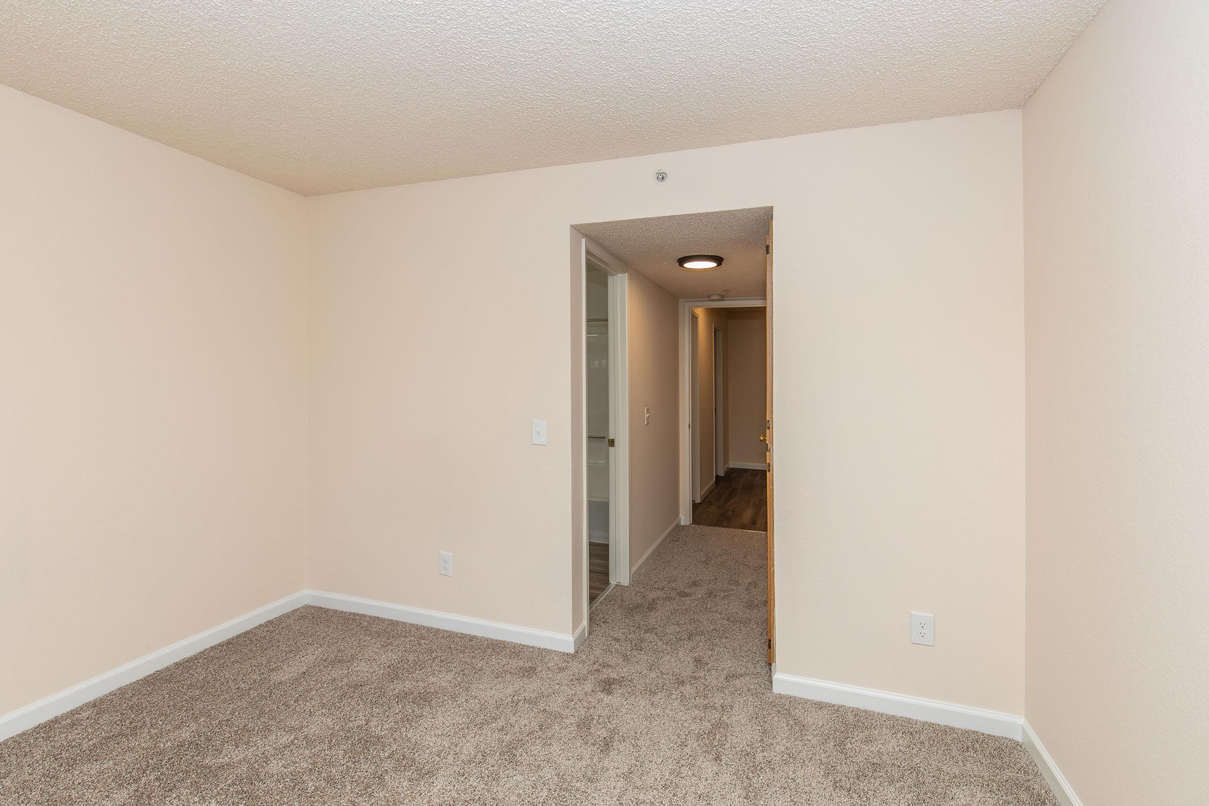 A small, empty room with light beige walls and plush carpet. The view includes an open doorway leading to a hallway. The room has a ceiling light fixture and is well-lit, showcasing a clean and minimal design. There are no furnishings present, emphasizing the spaciousness of the area.