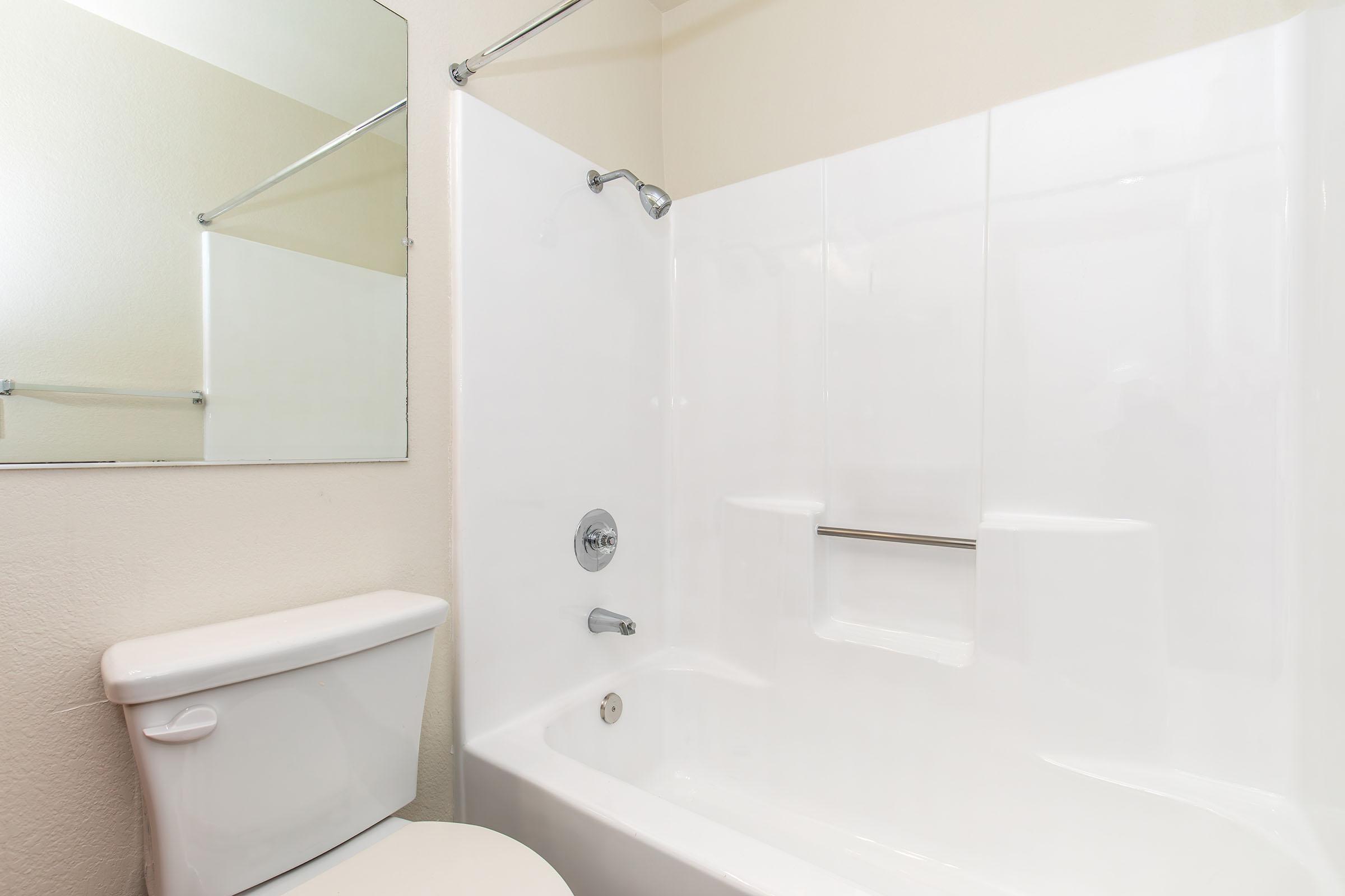 A clean, modern bathroom featuring a white bathtub and shower with a chrome faucet. A simple mirror is mounted on the wall, and a white toilet is positioned beside the bathtub. The walls are painted a light color, creating a bright and airy atmosphere.