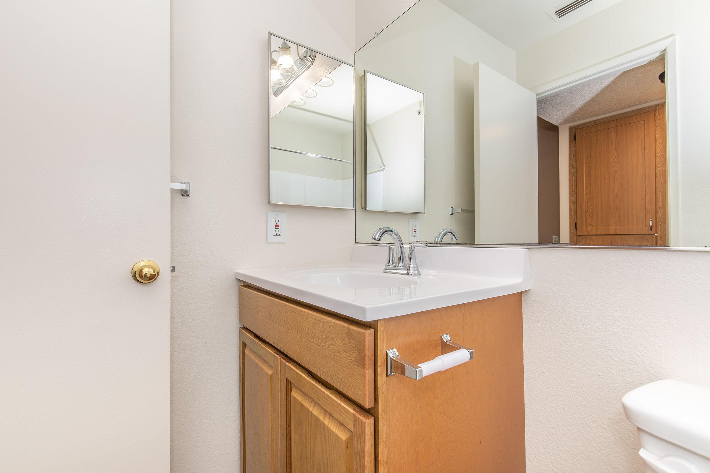 A clean and modern bathroom with a wooden vanity featuring a white countertop and two sinks. A large mirror hangs above the sinks. The room has light-colored walls and a door leading to a separate area. Natural light fills the space, creating a bright and inviting atmosphere.