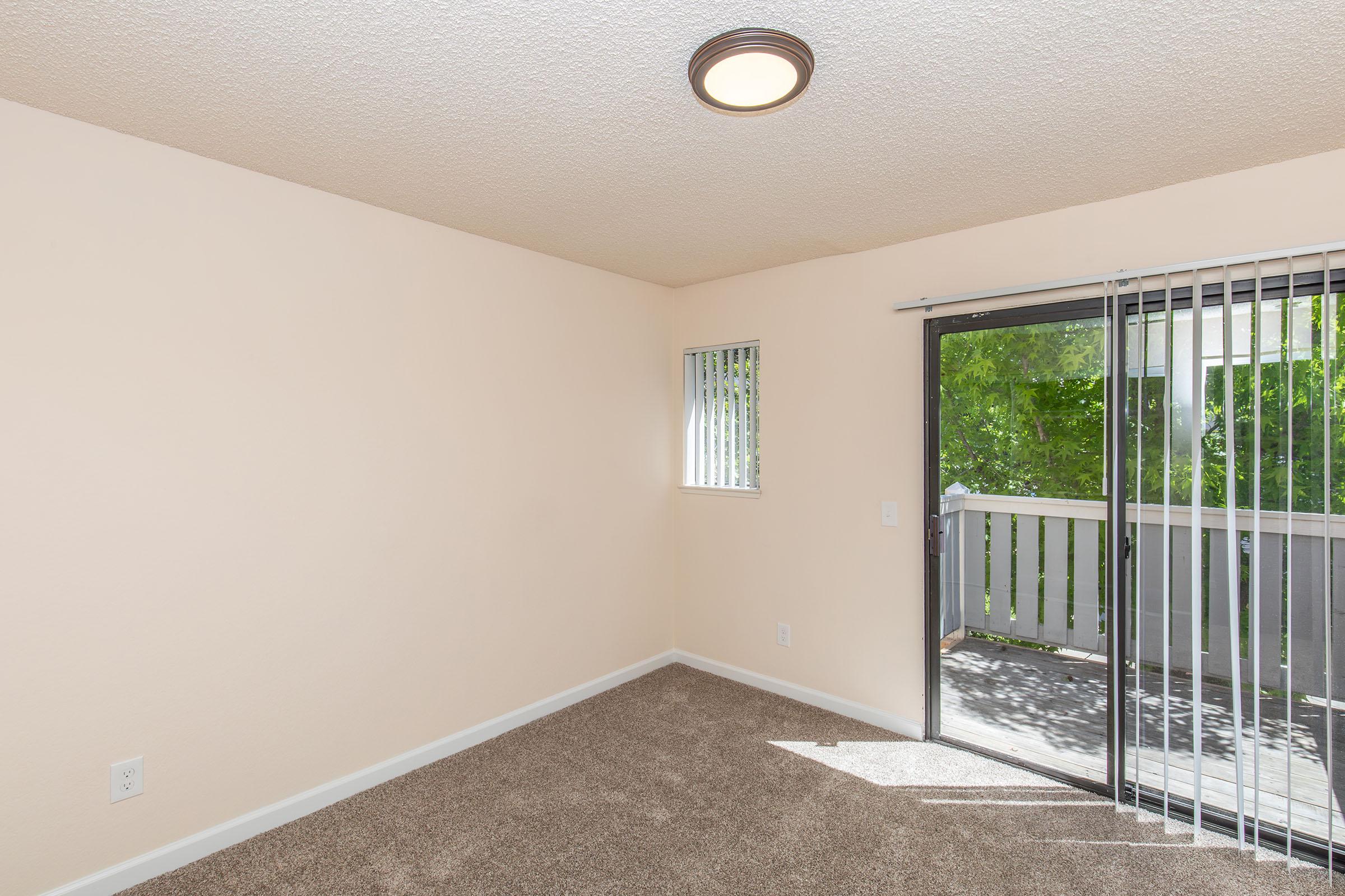 A well-lit, empty room with light beige walls and carpeted flooring. The room features a sliding glass door with vertical blinds leading to a balcony, and a small window with vertical blinds. A ceiling light fixture is visible, enhancing the bright appearance of the space.