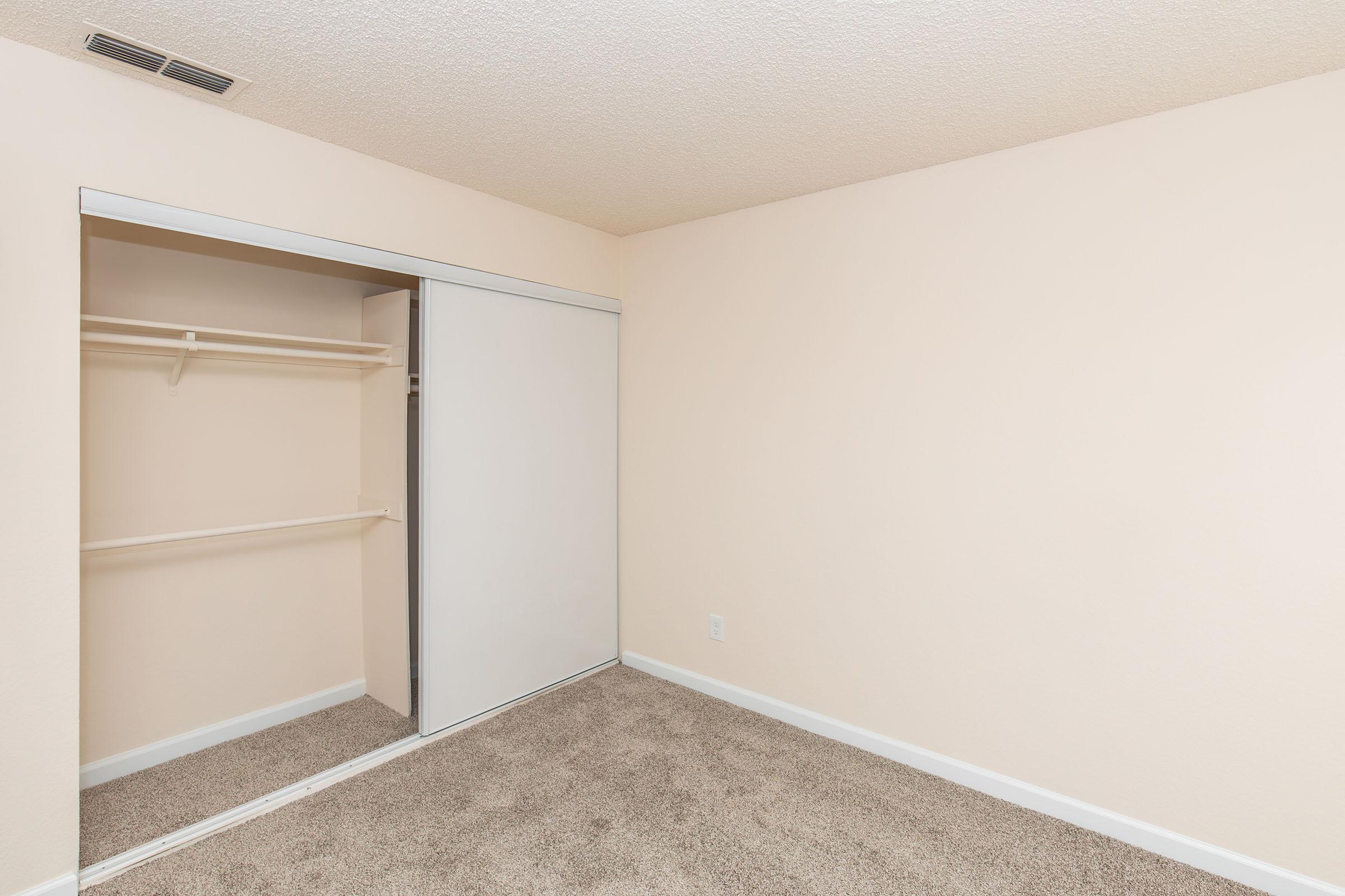 Interior view of a room with light beige walls and carpeted flooring. A sliding closet door is visible on the left, with a single shelf inside. The room appears empty and well-lit, offering a clean, minimalist aesthetic.