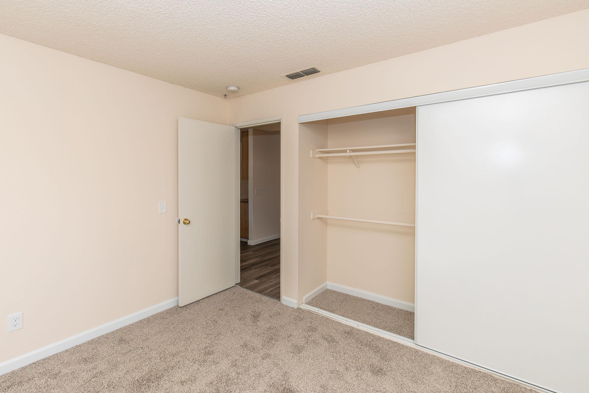 A small, empty room featuring light beige walls and a plush carpet. A door is partially open, revealing a hallway leading to another area. On one side, there is a closet with white sliding doors, with a rod for hanging clothes visible inside. The room has a bright and clean appearance, suitable for various uses.