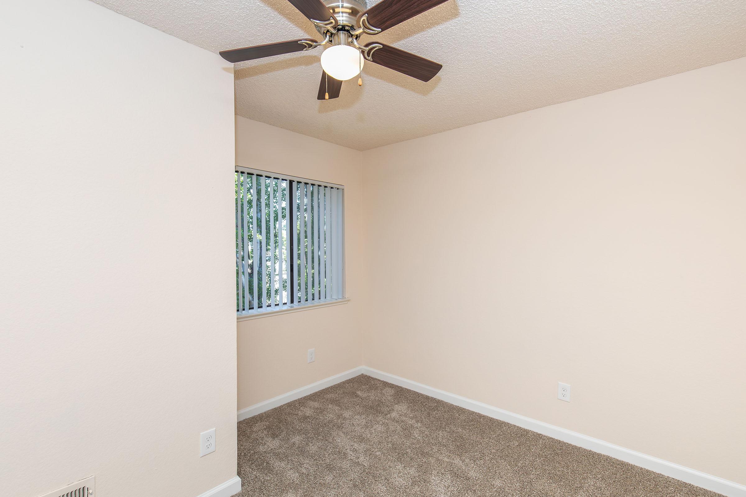 A small, empty room with beige walls and carpet. It features a ceiling fan and a window with vertical blinds that lets in natural light. The room has a simple layout with no furnishings, creating a spacious and airy feel.