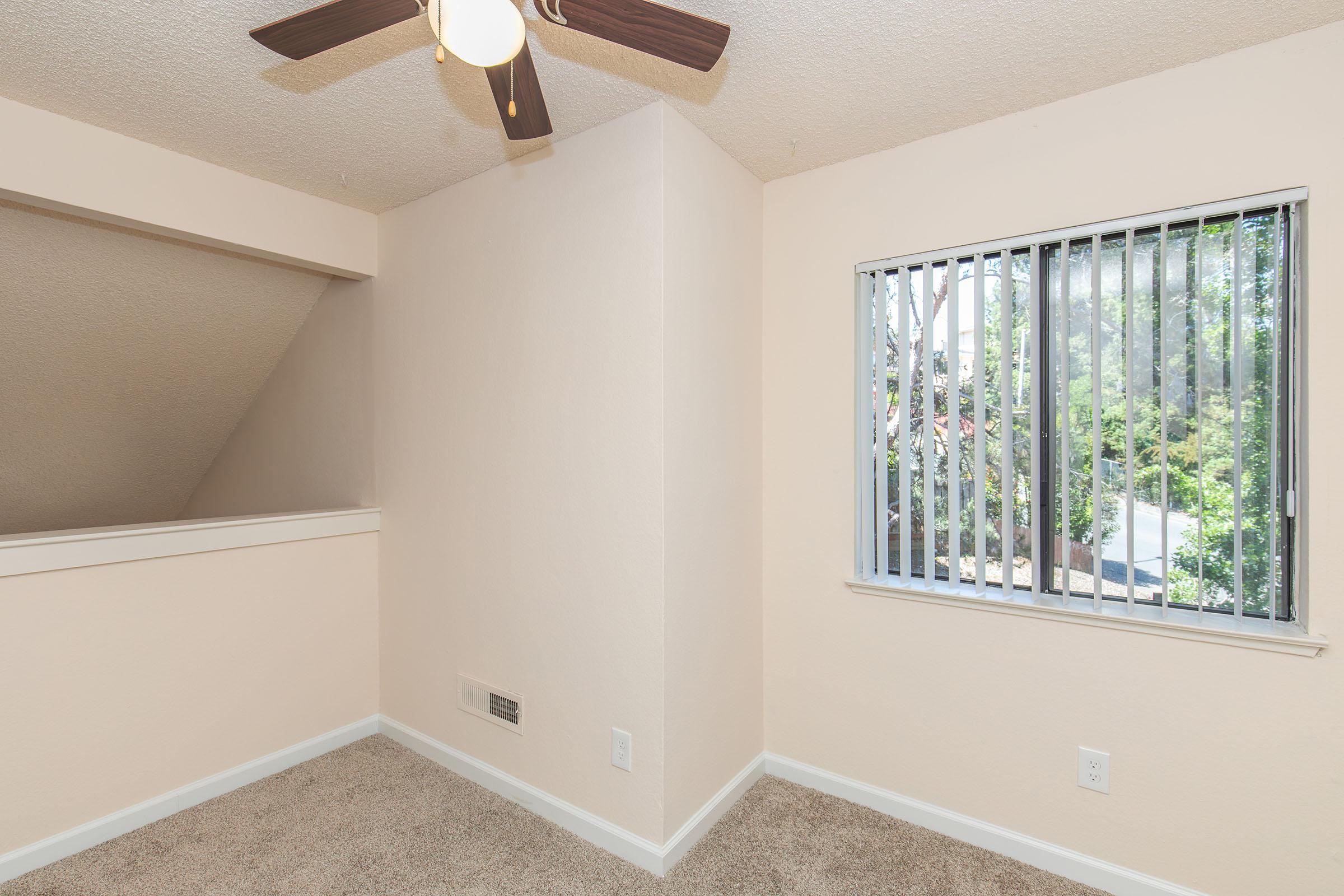 A cozy, light-colored room featuring beige walls and carpet, with a ceiling fan and a window adorned with vertical blinds, looking out to a green area outside. The room has a simple, clean design, ideal for relaxation or a small workspace.
