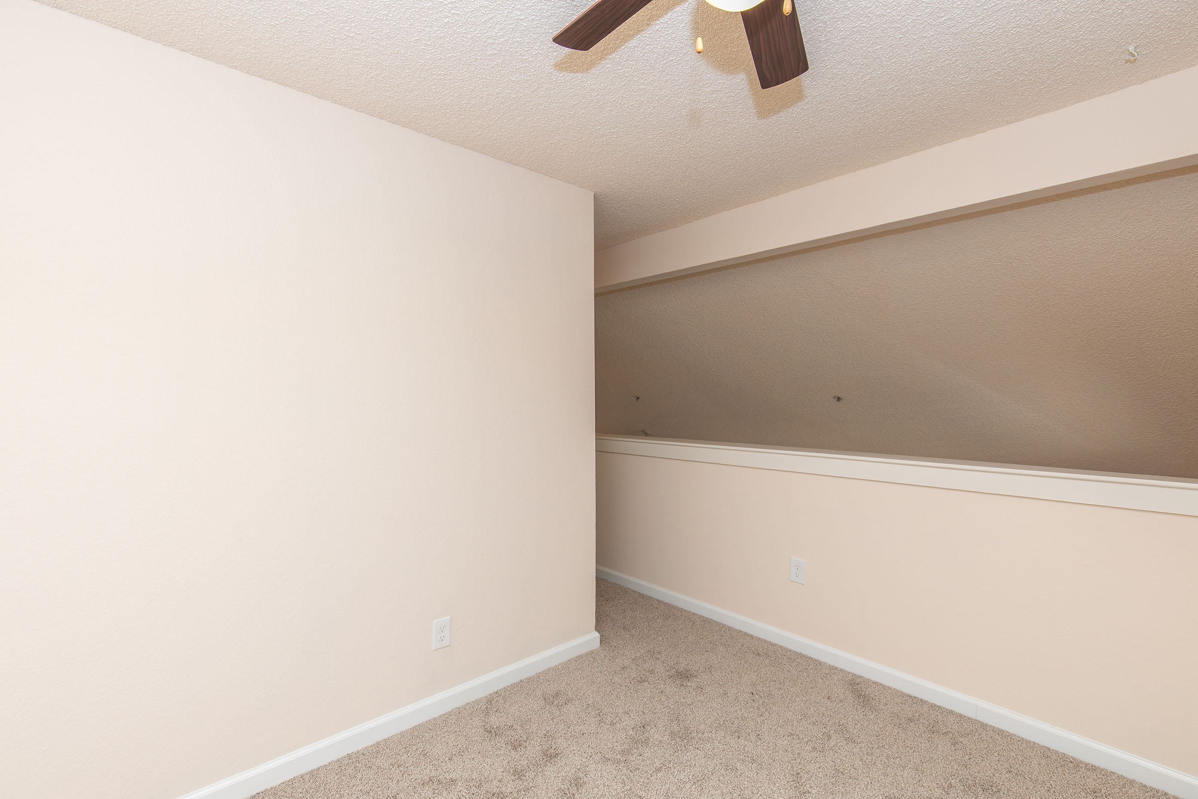 A simple room featuring beige walls and carpeting, with a slanted ceiling. A light-colored handrail is visible along the wall, and a ceiling fan is installed. The overall space appears well-lit and empty, suggesting it could be used as an office or small bedroom.