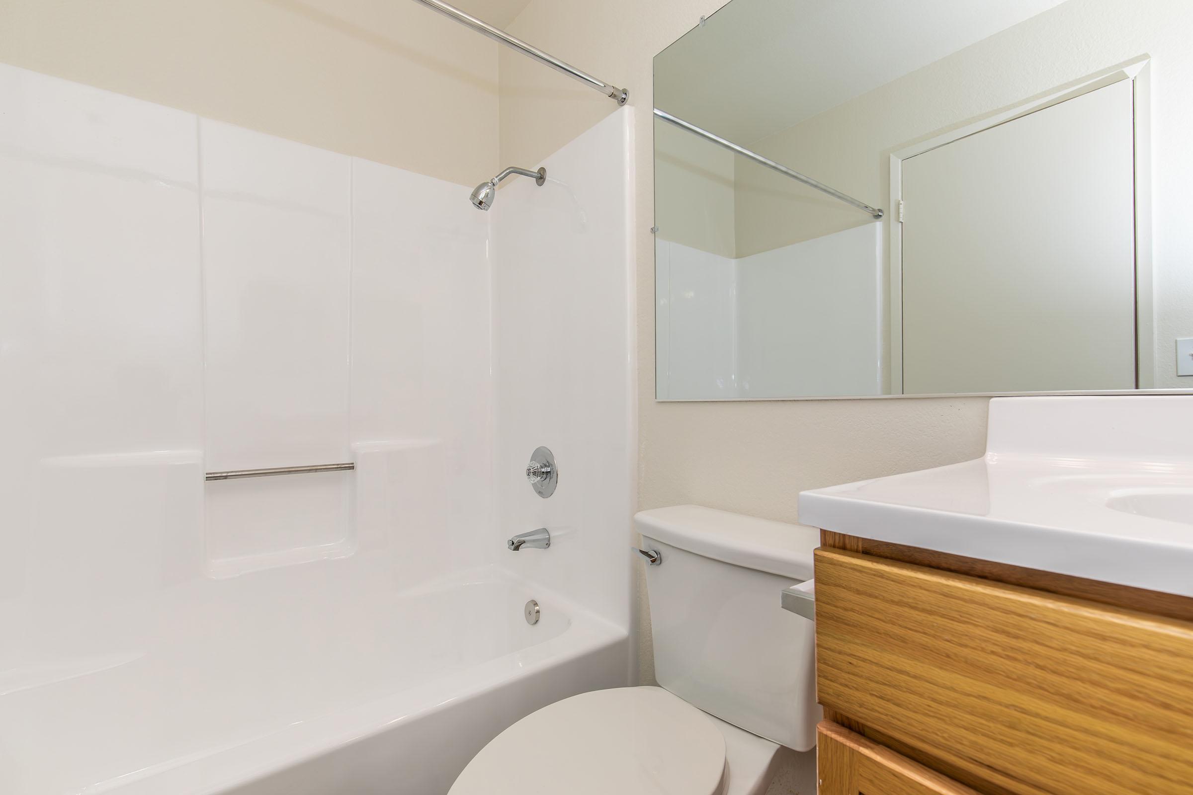 A clean, modern bathroom featuring a white bathtub with a showerhead, a mirror above the sink, a wooden cabinet under the sink, and a toilet. The walls are painted a light color, and the overall space appears bright and tidy, emphasizing a minimalist design.