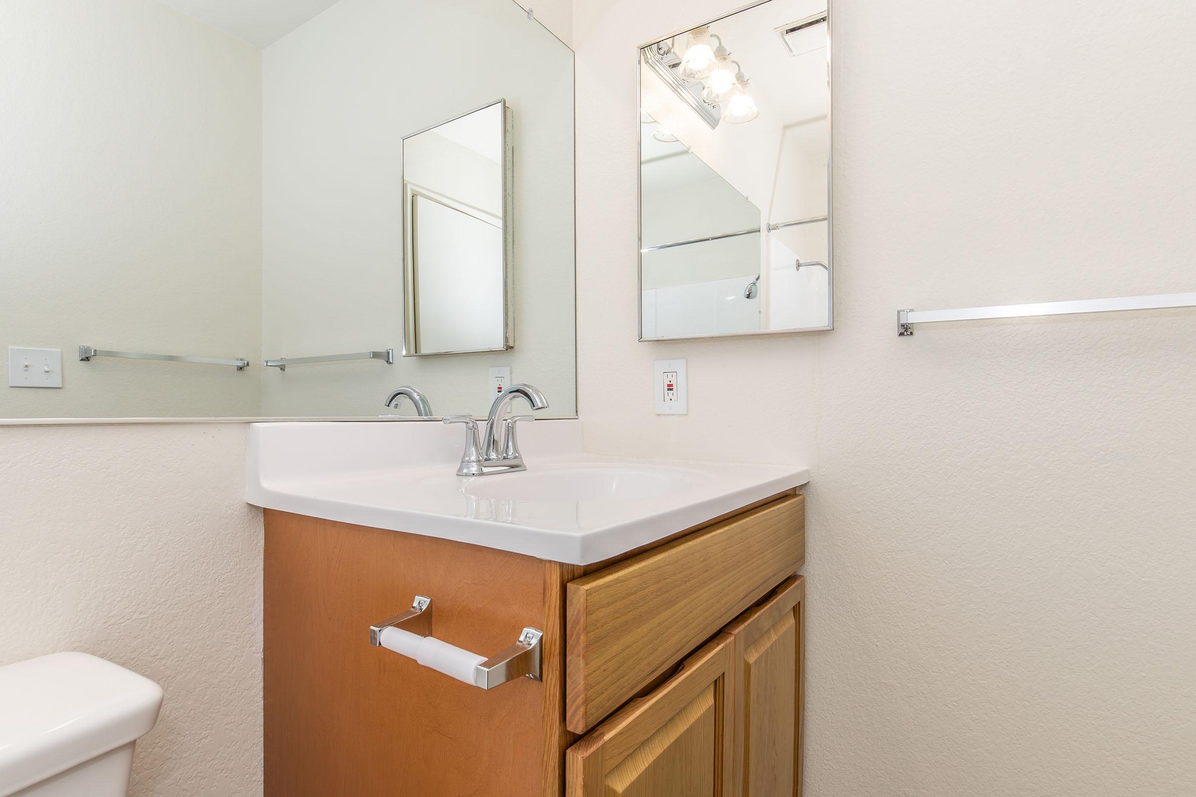 A clean and modern bathroom featuring a double sink vanity with wooden cabinetry, a large mirror above the sink, and a bathroom wall with light-colored paint. The space is well-lit with overhead lighting and showcases a simple, functional design.