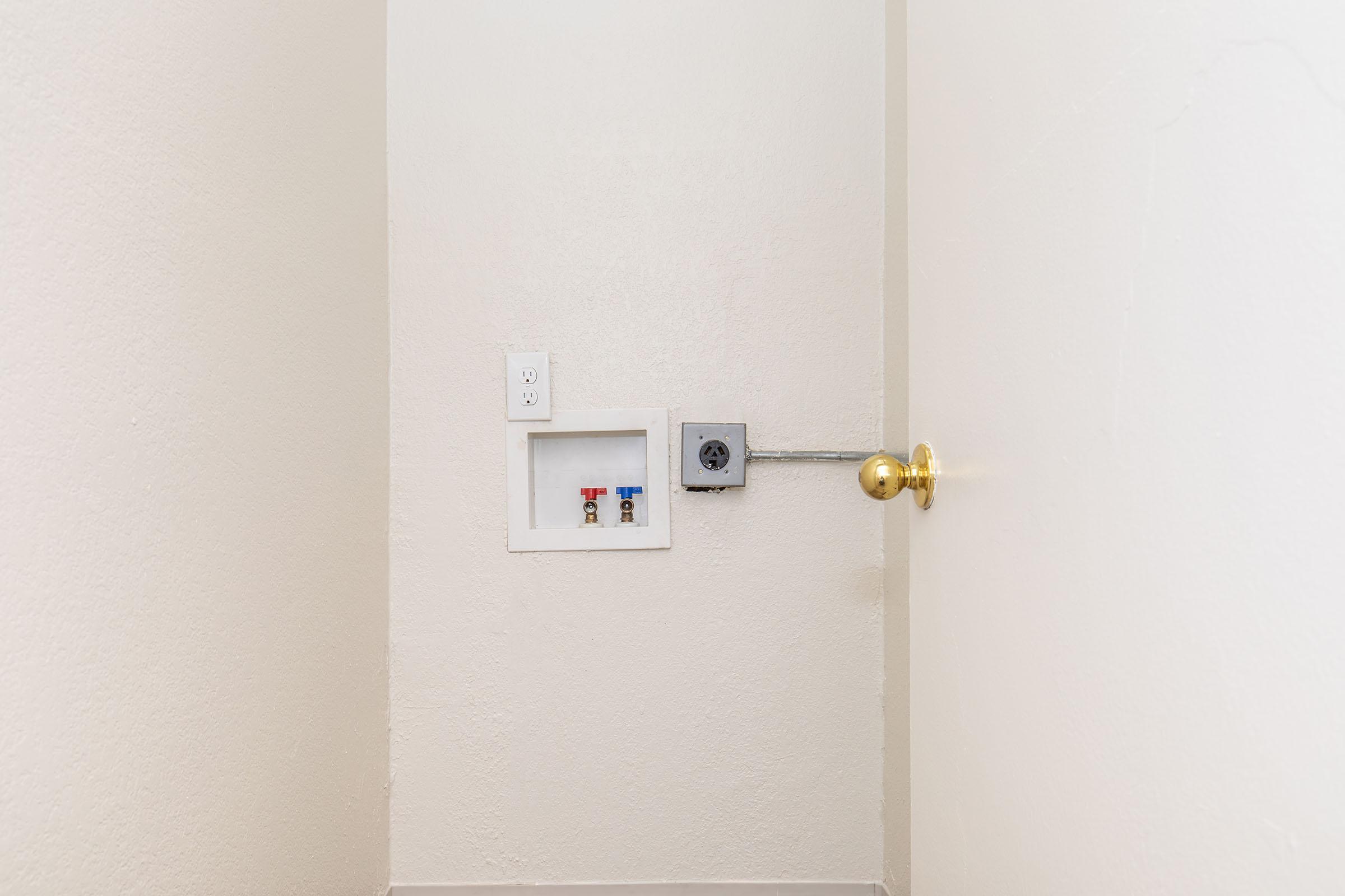 A corner of a room with light-colored walls, featuring a small electrical panel or outlet box with colored toggle switches and a power outlet on the left side. A round doorknob is visible on the right side, leading to another space. The flooring is light and minimalistic.