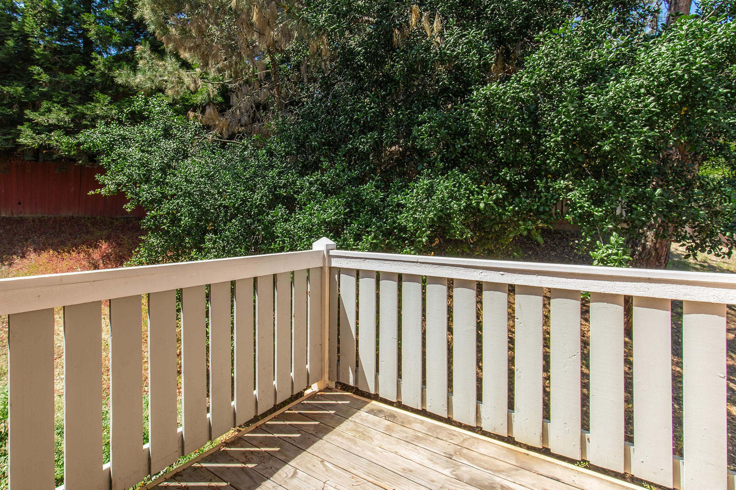 A wooden deck railing with a view of lush green trees in the background, casting shadows on the wooden floor. The scene is peaceful and natural, indicating a quiet outdoor space.