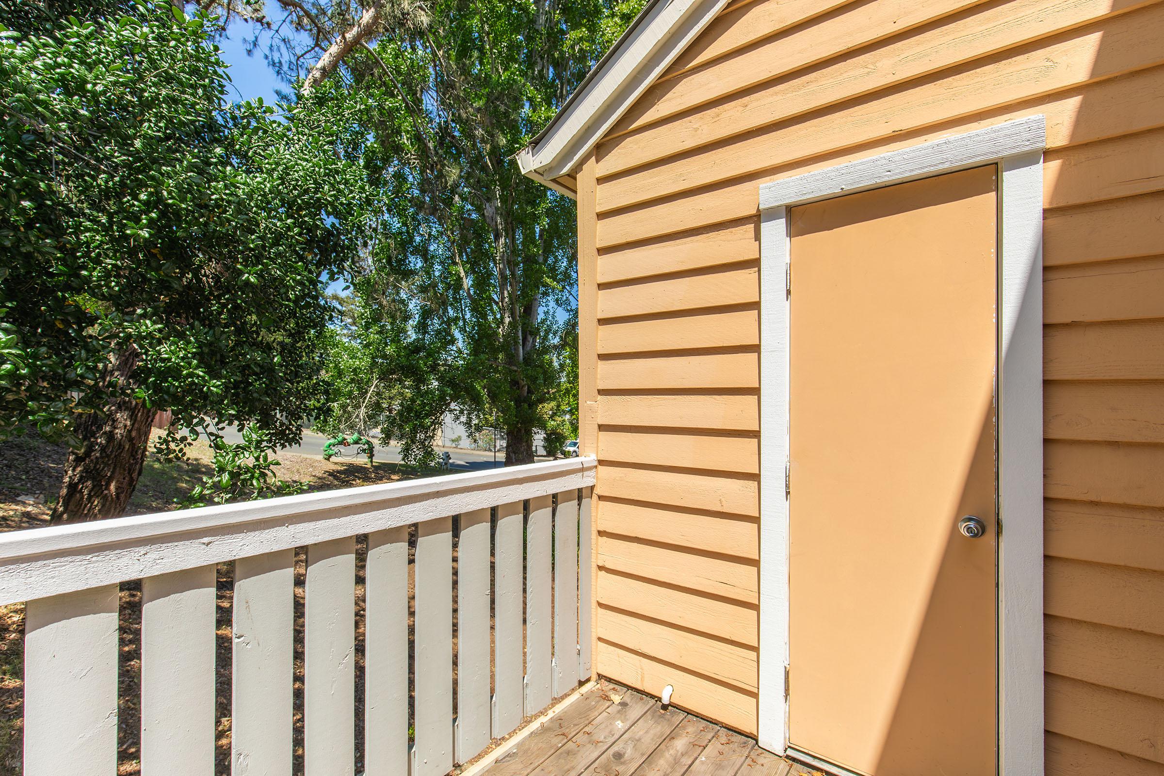 A close-up view of the corner of a wooden house featuring a peach-colored door on a light yellow wall. A white railing surrounds a small porch area, and greenery from nearby trees is visible in the background under a clear blue sky.
