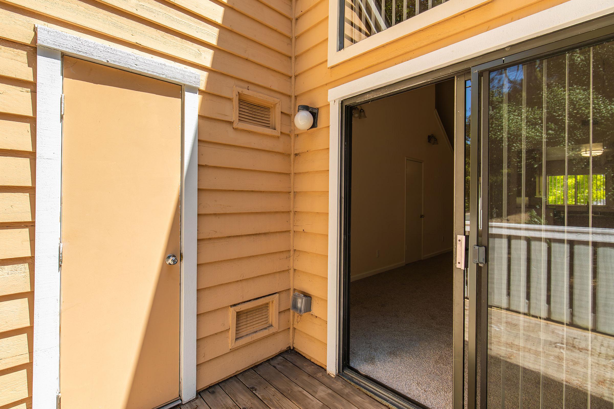 A sunny exterior view of a light orange house showing a closed door and an open sliding glass door leading into a well-lit, empty room with carpet. The room has a window with a view of greenery outside. The walls are painted in a soft beige color, creating a warm atmosphere.
