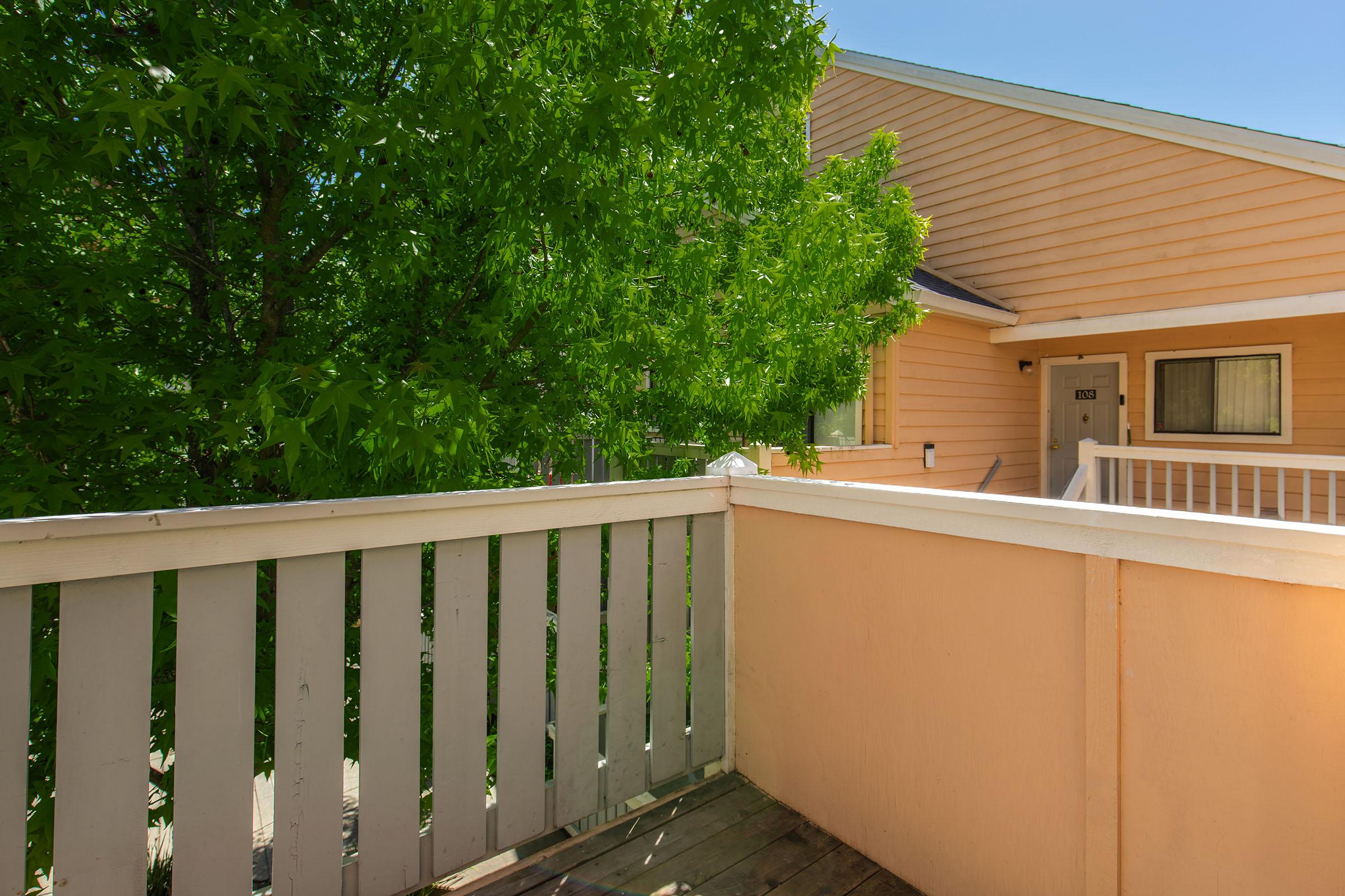 A wooden balcony with a white railing overlooks a green tree. In the background, a peach-colored building features a door and a window. Bright sunlight illuminates the scene, creating a serene atmosphere.