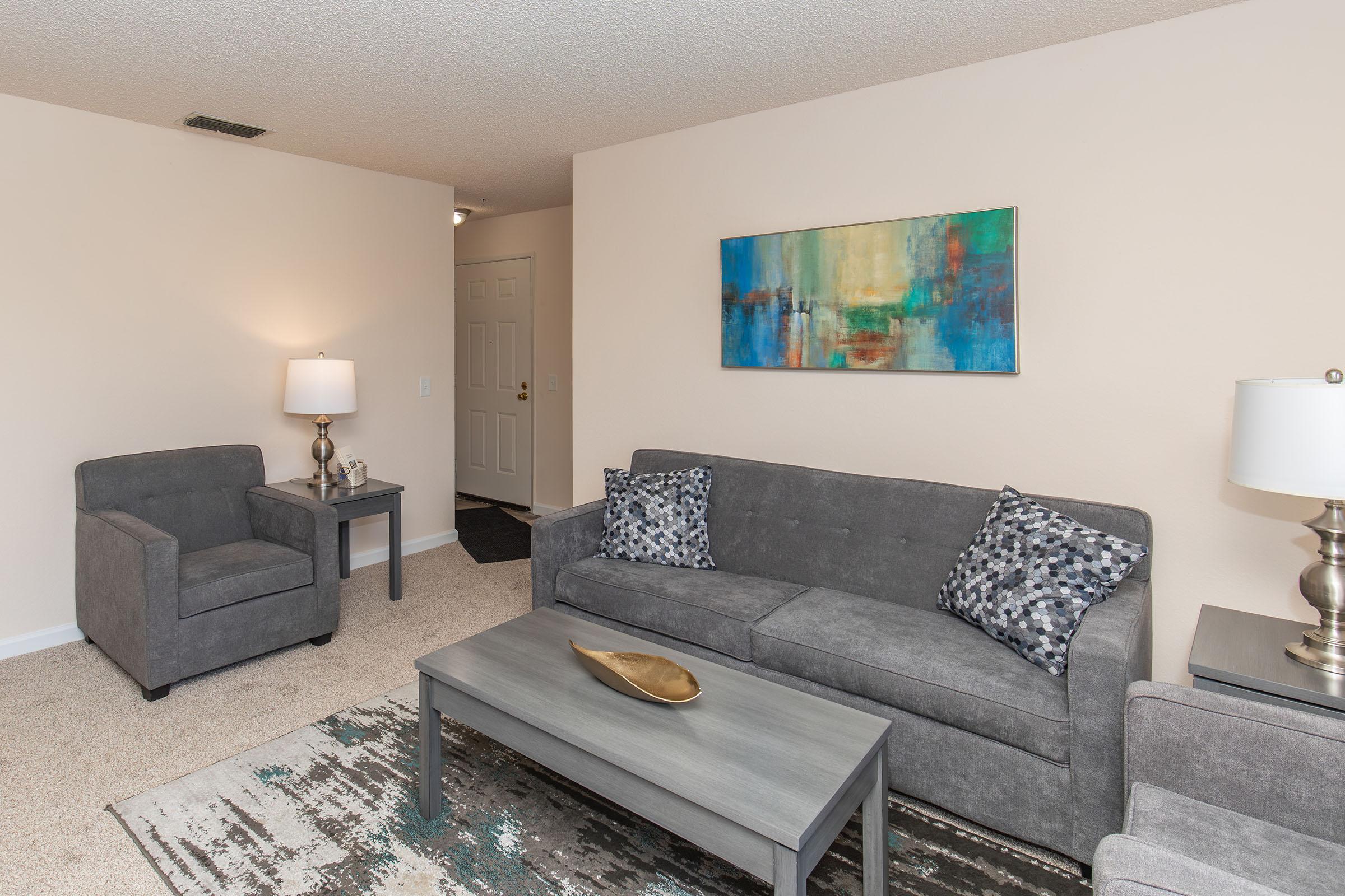 A cozy living room featuring a gray sofa with decorative pillows, an armchair, a coffee table with a decorative bowl, and two lamps. A colorful abstract painting hangs on the wall, and the room has neutral-colored walls and carpet, offering a warm and inviting atmosphere.