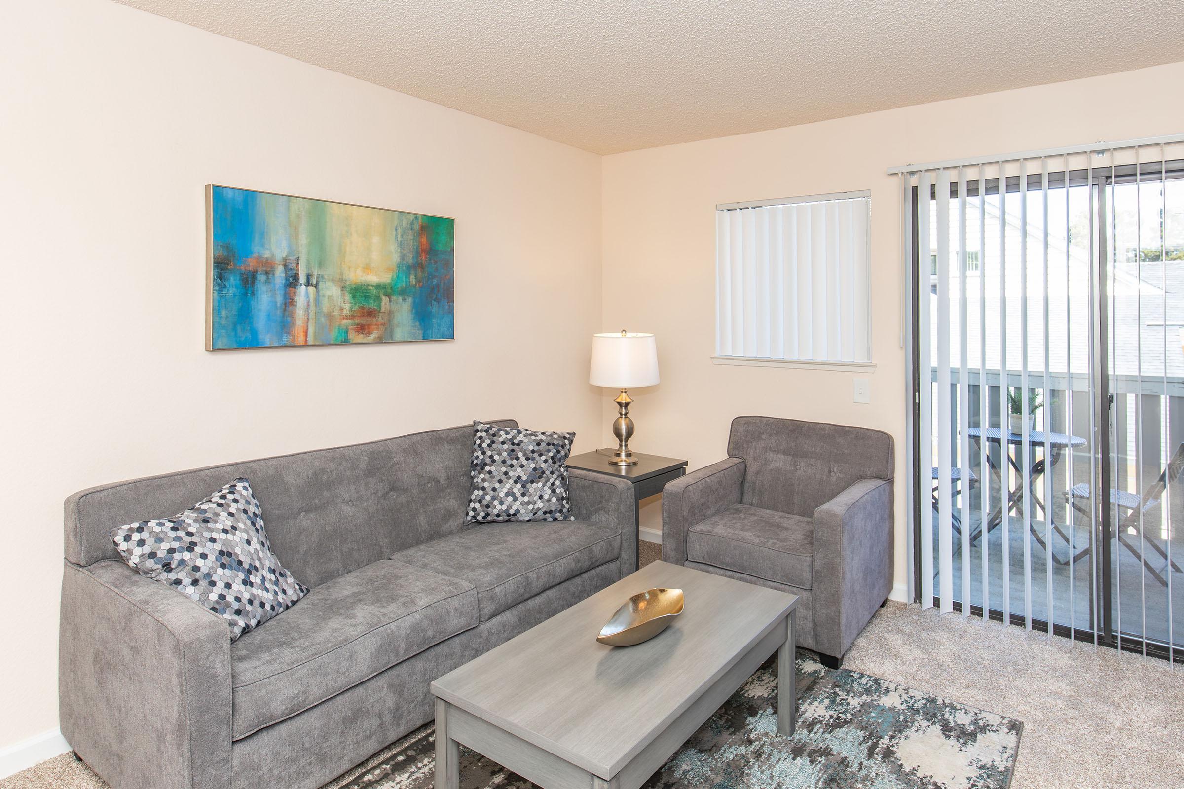 A modern living room featuring a gray couch with decorative pillows, a matching armchair, a light gray coffee table, and an abstract painting on the wall. Natural light flows in through vertical blinds, and there is a small balcony visible in the background. The space is stylishly decorated and inviting.
