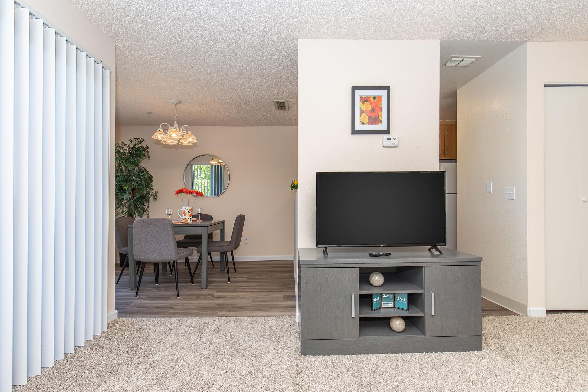A living room with a modern TV stand and a television. In the background, there's a dining area with a round table set for a meal, featuring a vase of flowers. The walls are painted light colors, and sliding glass doors provide natural light. The floor is covered with carpet, and there are plants for decoration.