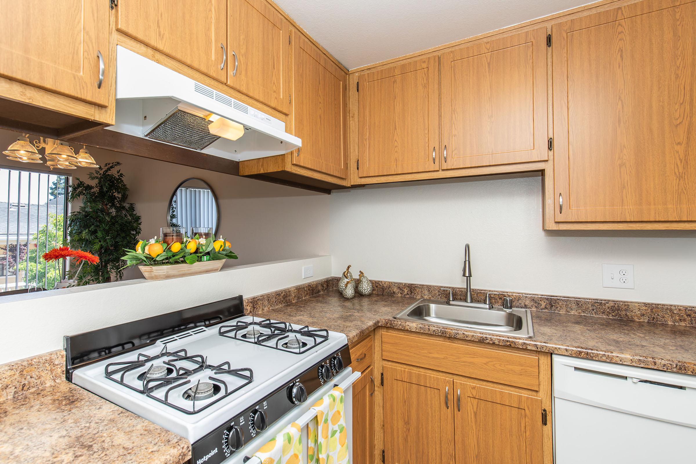 A modern kitchen with wooden cabinets, a gas stove, and a stainless steel sink. There is a window with a circular mirror and a bowl of colorful fruit on the counter. The countertop is light brown with a decorative towel hanging from the stove. The overall design is bright and inviting.