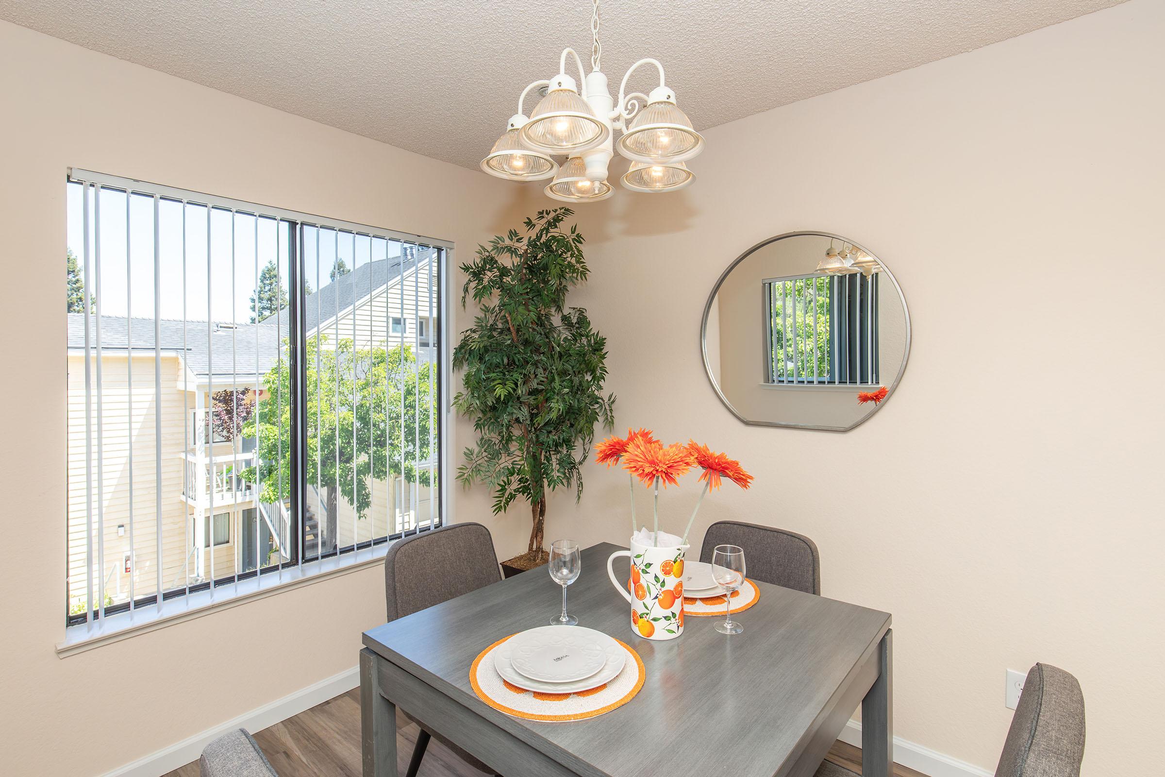 A bright dining area featuring a modern table with gray chairs, adorned with a floral centerpiece. A stylish chandelier hangs above, and natural light enters through a large window with vertical blinds. A round mirror reflects the space, complemented by greenery nearby.