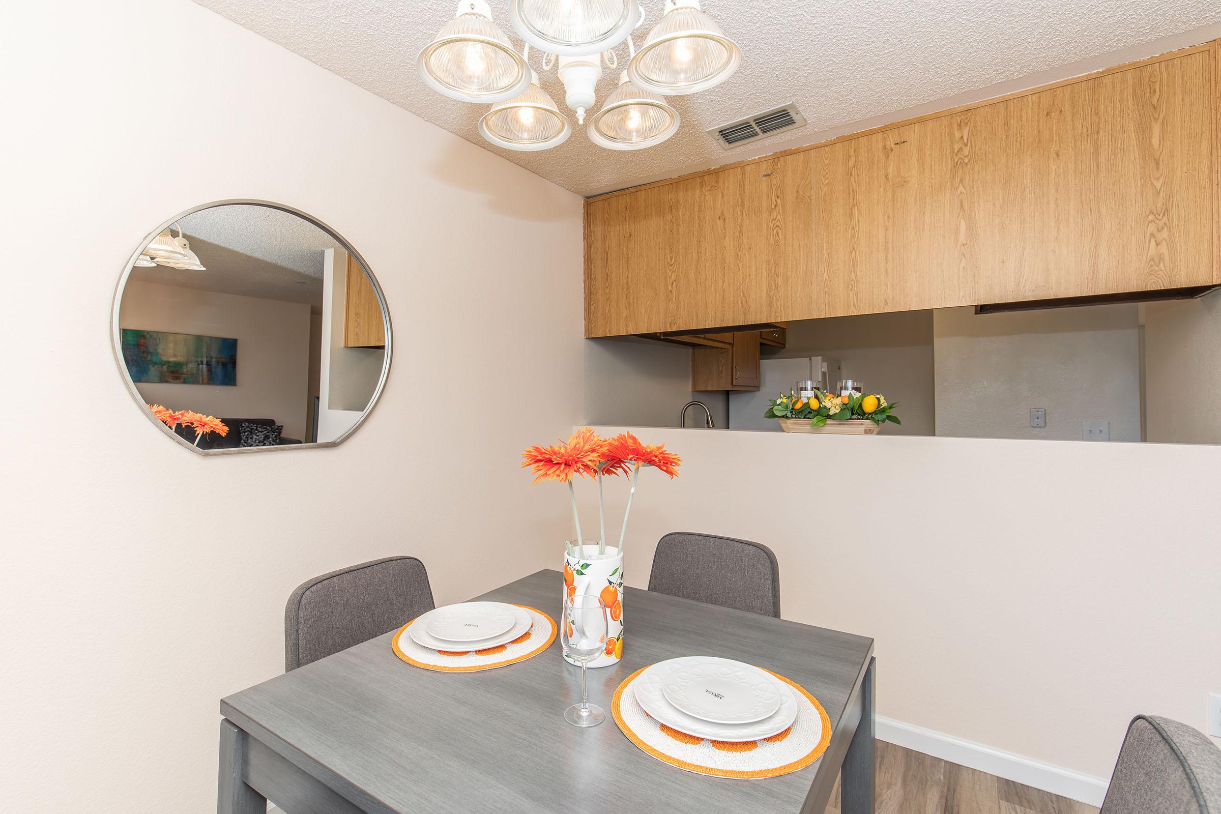 A modern dining area featuring a round mirror on the wall, a table set for two with plates and a vase of orange flowers. The kitchen area is visible in the background with wooden cabinetry and a floral arrangement. Soft lighting is provided by a stylish chandelier above the table.