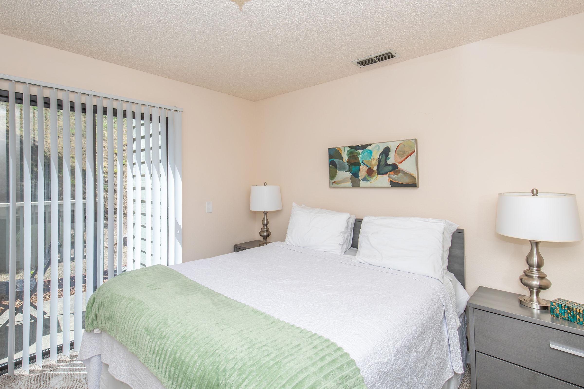 A cozy bedroom featuring a queen-sized bed with white linens and a green throw blanket. On either side of the bed are modern silver lamps on matching nightstands. A colorful abstract painting hangs above the bed. Natural light enters through vertical blinds, creating a bright and inviting atmosphere.