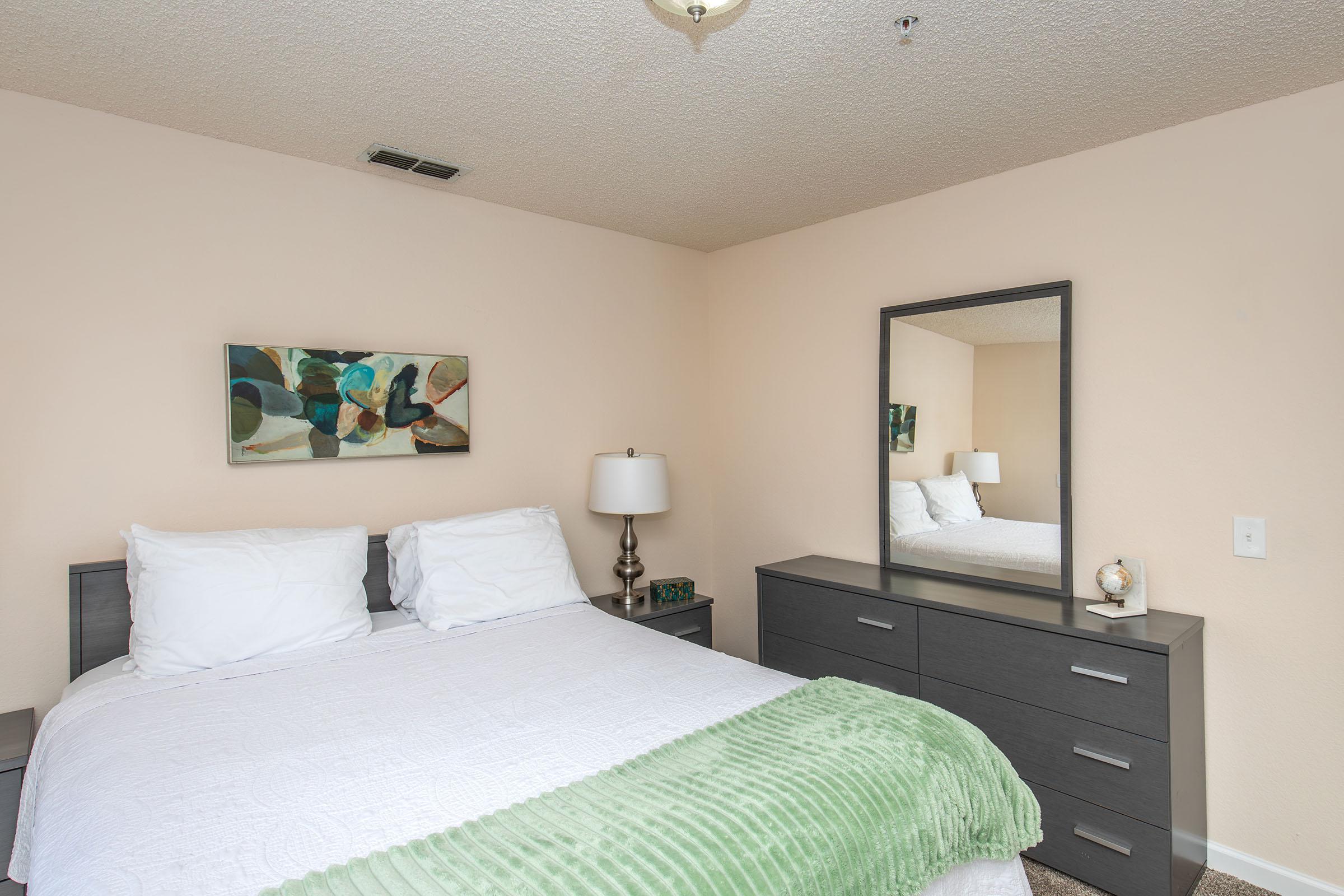 A cozy bedroom featuring a bed with white bedding and green accents, a modern dresser, a table lamp, and a mirror. Soft pastel walls create a warm atmosphere, and abstract art hangs above the bed, adding a touch of color. Natural light enhances the inviting ambiance.