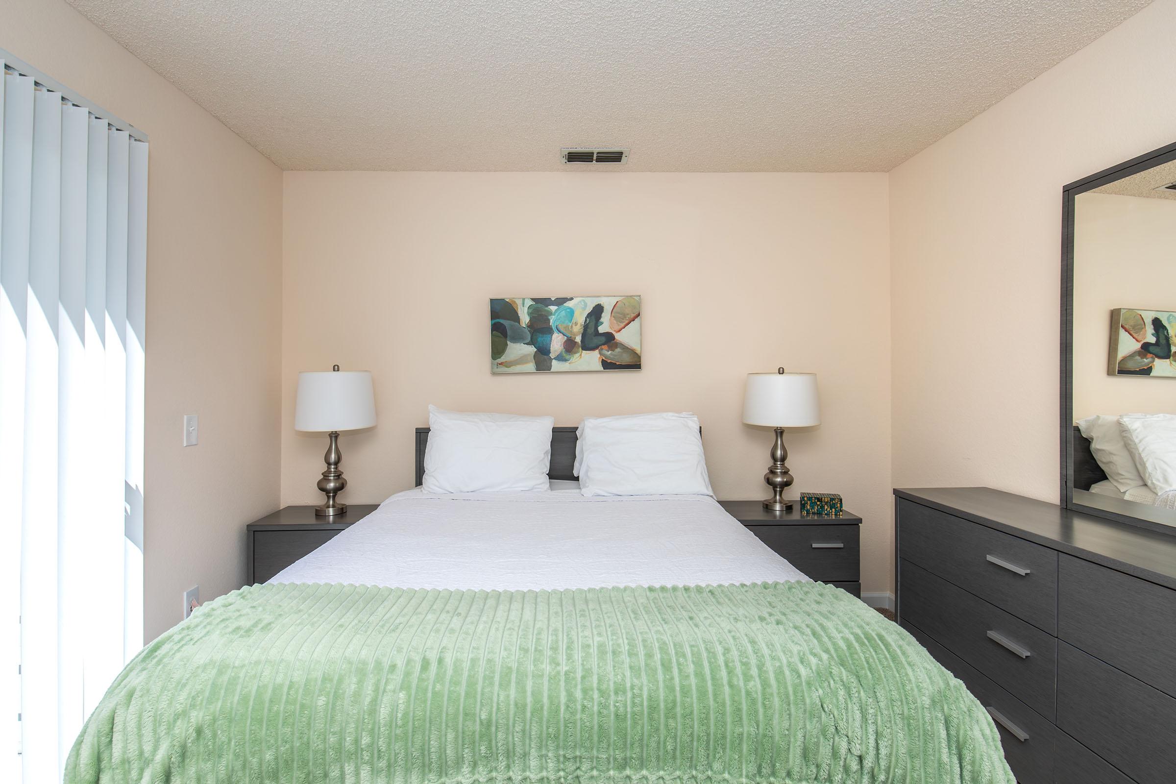 A cozy bedroom featuring a queen-sized bed with white bedding and a green throw blanket. Two bedside lamps sit on wooden nightstands. A mirror reflects the room, and a piece of abstract art hangs on the wall above the bed. Natural light streams through vertical blinds, creating a welcoming atmosphere.