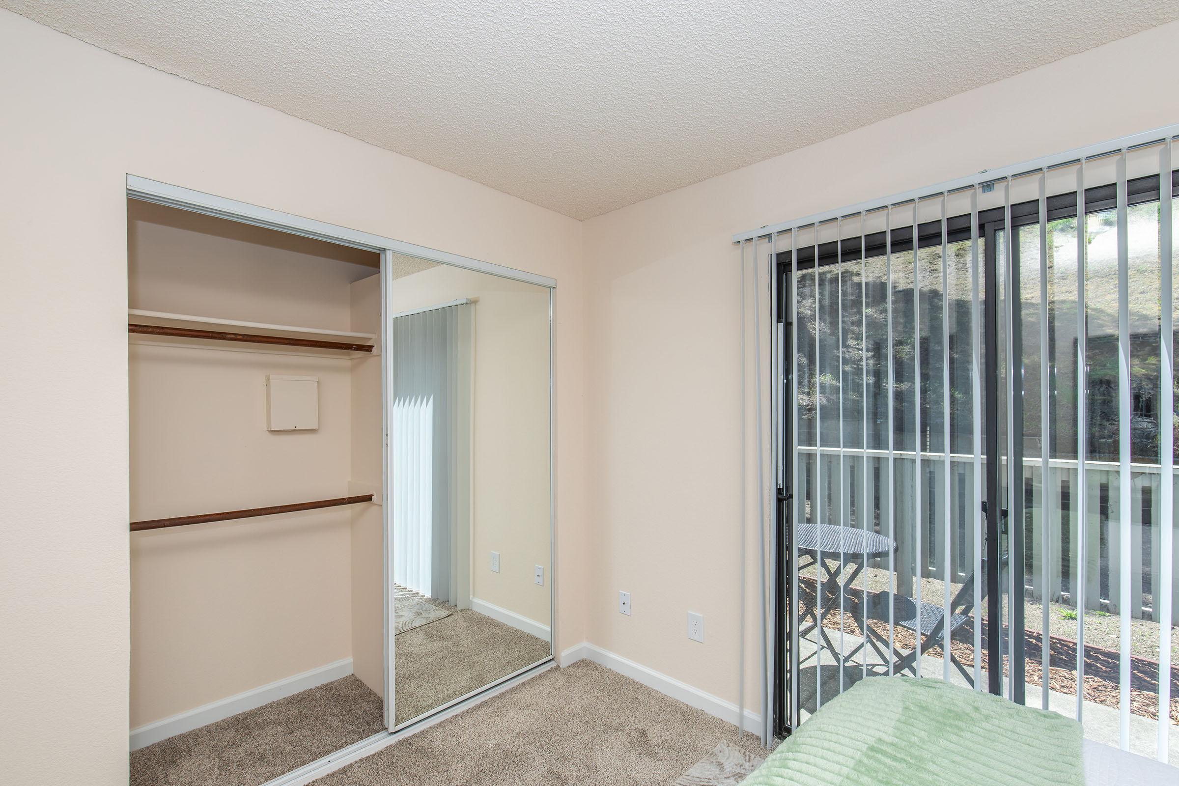 Interior view of a room with a mirror closet door and a small table and chairs visible outside through sliding glass doors. The walls are light-colored and the carpet is beige, creating a warm and inviting atmosphere. Natural light streams in, highlighting the spacious feel of the room.