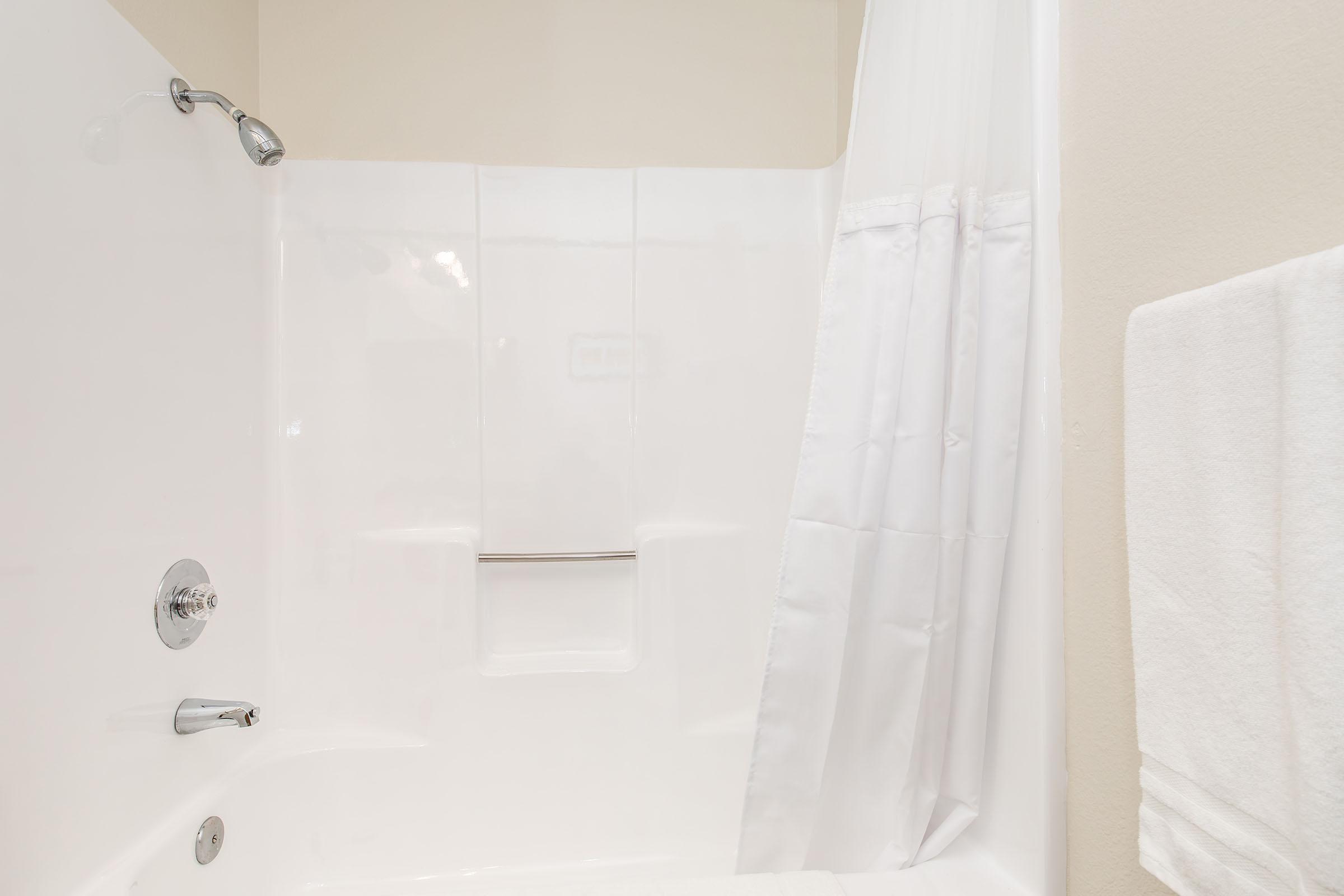 A clean and bright bathroom featuring a white bathtub with a showerhead, a smooth white tub surround, a folded white towel hanging on a towel rack, and a white shower curtain neatly drawn to one side, showcasing a simple and functional design.