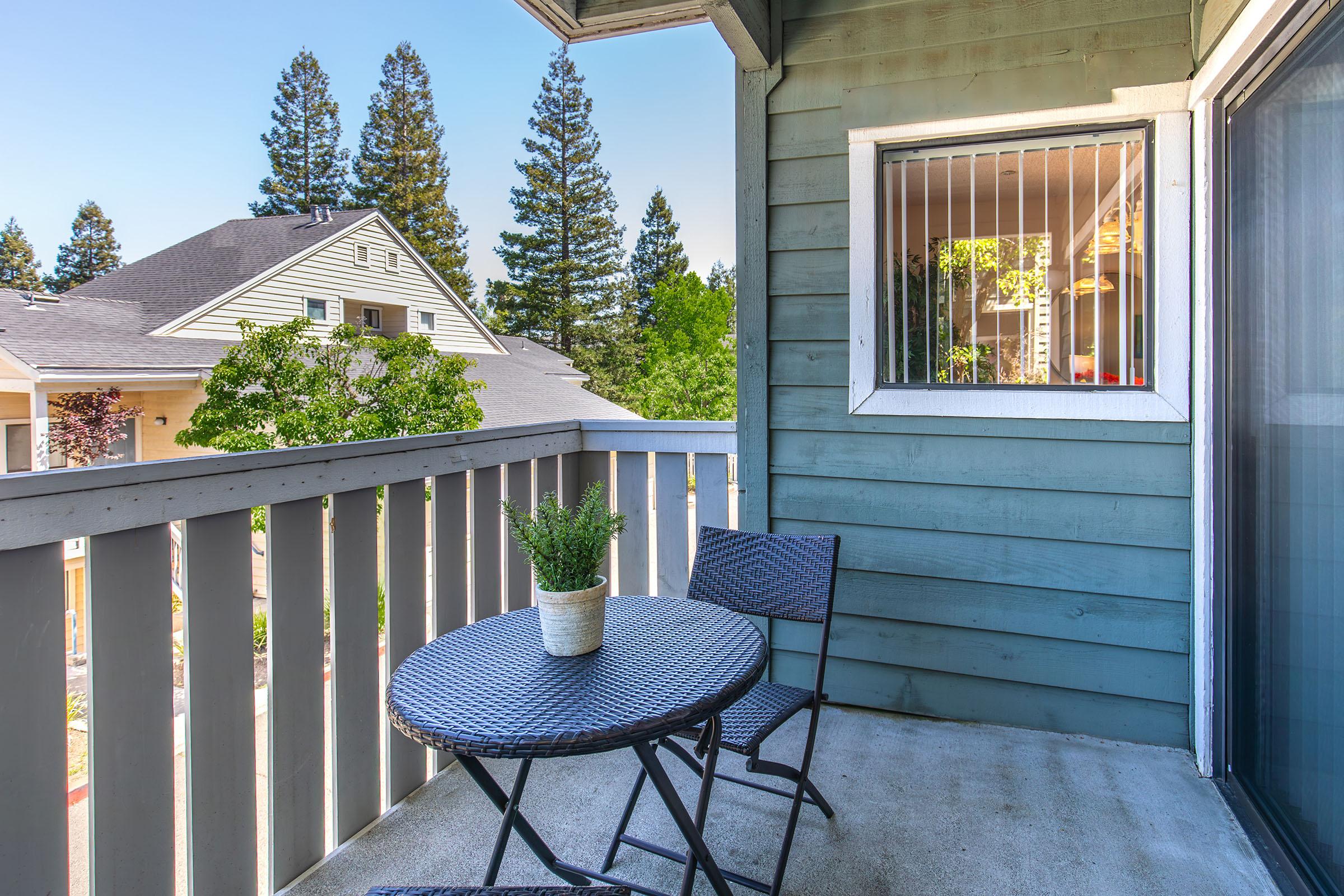 A cozy balcony featuring a small round table and two chairs. A potted plant sits on the table. In the background, there are trees and another building visible, indicating a serene environment. The blue walls of the balcony add a calming touch to the outdoor space.