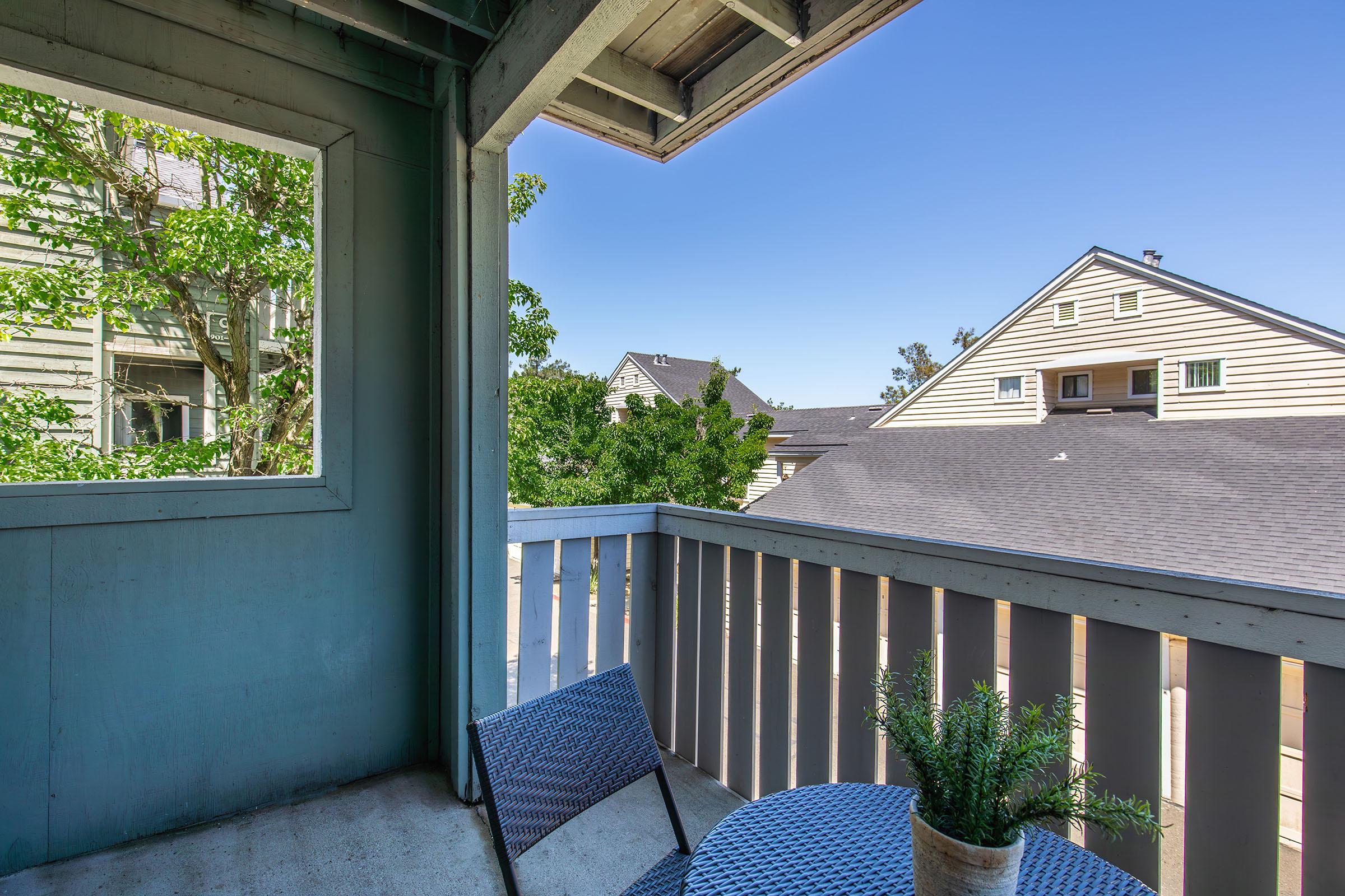A cozy balcony featuring a small round table and chairs, with a potted plant on the table. The view includes neighboring buildings and trees, under a clear blue sky, creating a serene outdoor space for relaxation.