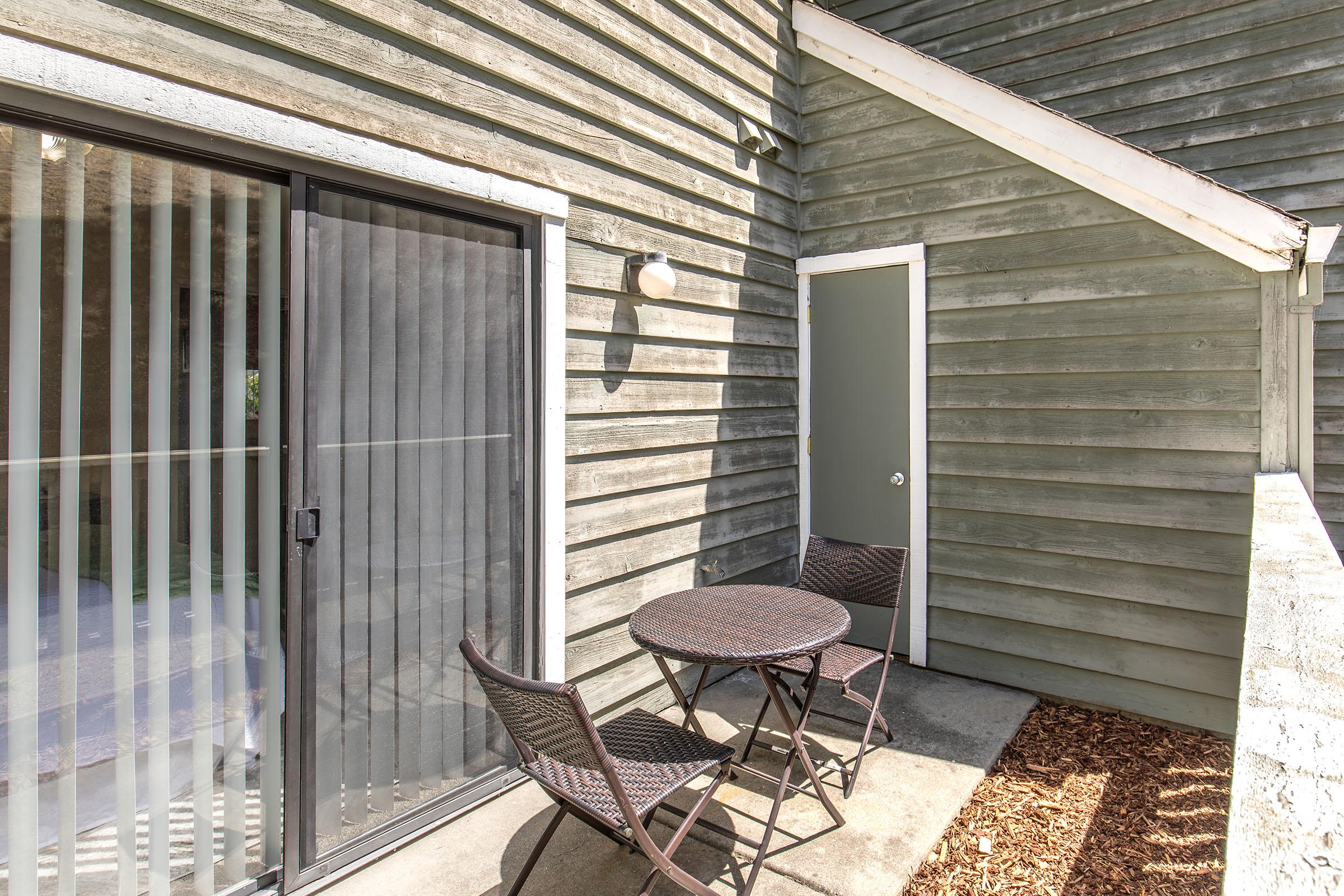 A small outdoor patio featuring a round table and two chairs, with a sliding glass door that leads inside. The walls are made of weathered wood, and the area is surrounded by a light gravel or mulch surface. The patio is illuminated by a wall-mounted light fixture.