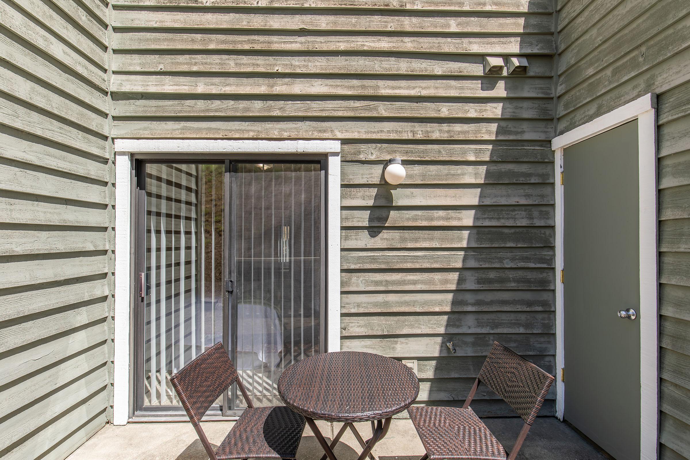 A small outdoor patio featuring a round table and two chairs. There's a sliding glass door that leads inside, flanked by wooden siding painted in a muted green color. A light fixture is mounted on the wall next to the door. Sunlight casts soft shadows on the surface.