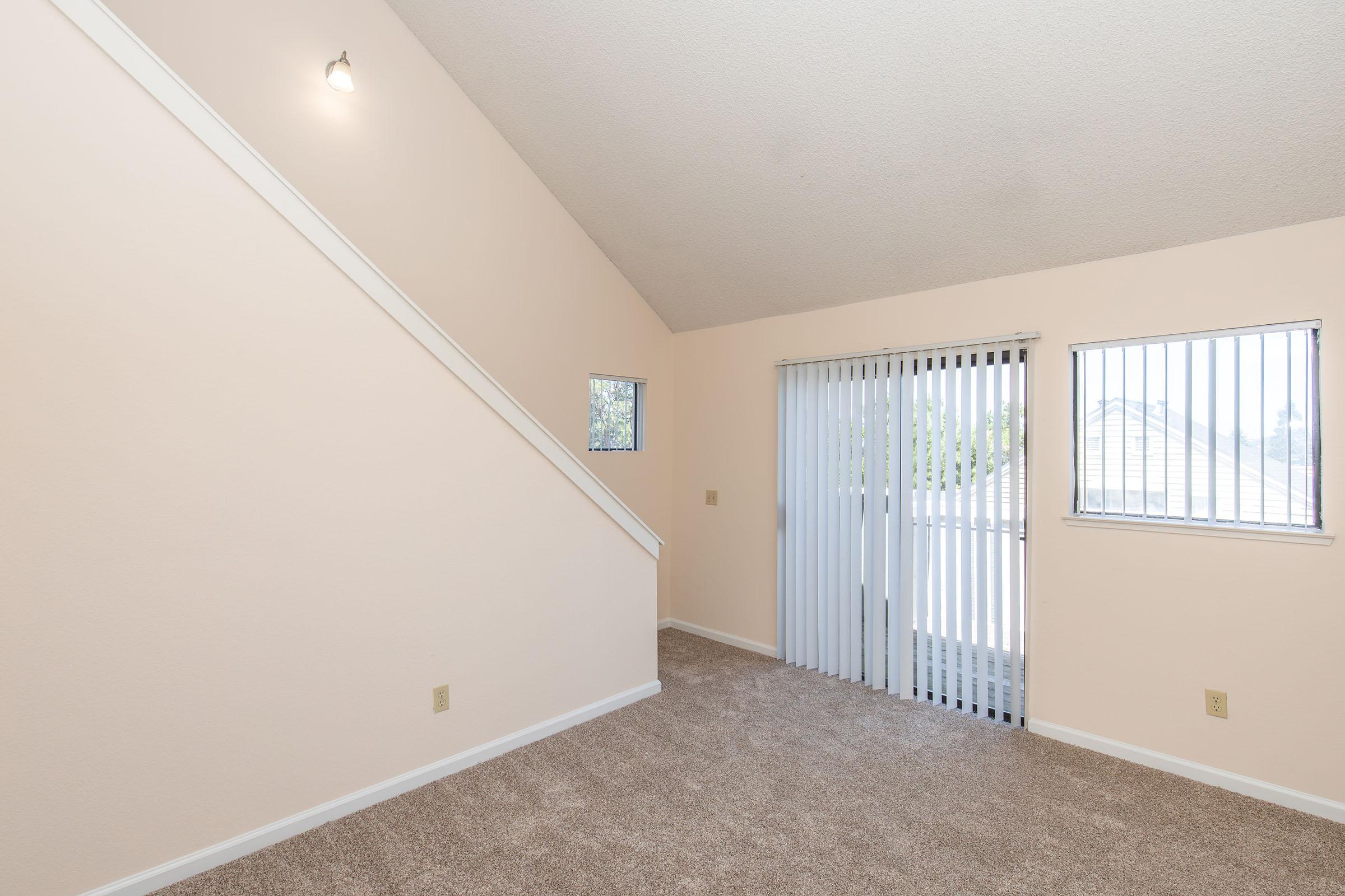 A spacious, empty room featuring light beige walls and plush carpeting. A staircase is visible on the left side, and large sliding glass doors with vertical blinds provide natural light and access to a small outdoor area. Two windows are on the right side, enhancing the airy feel of the space.