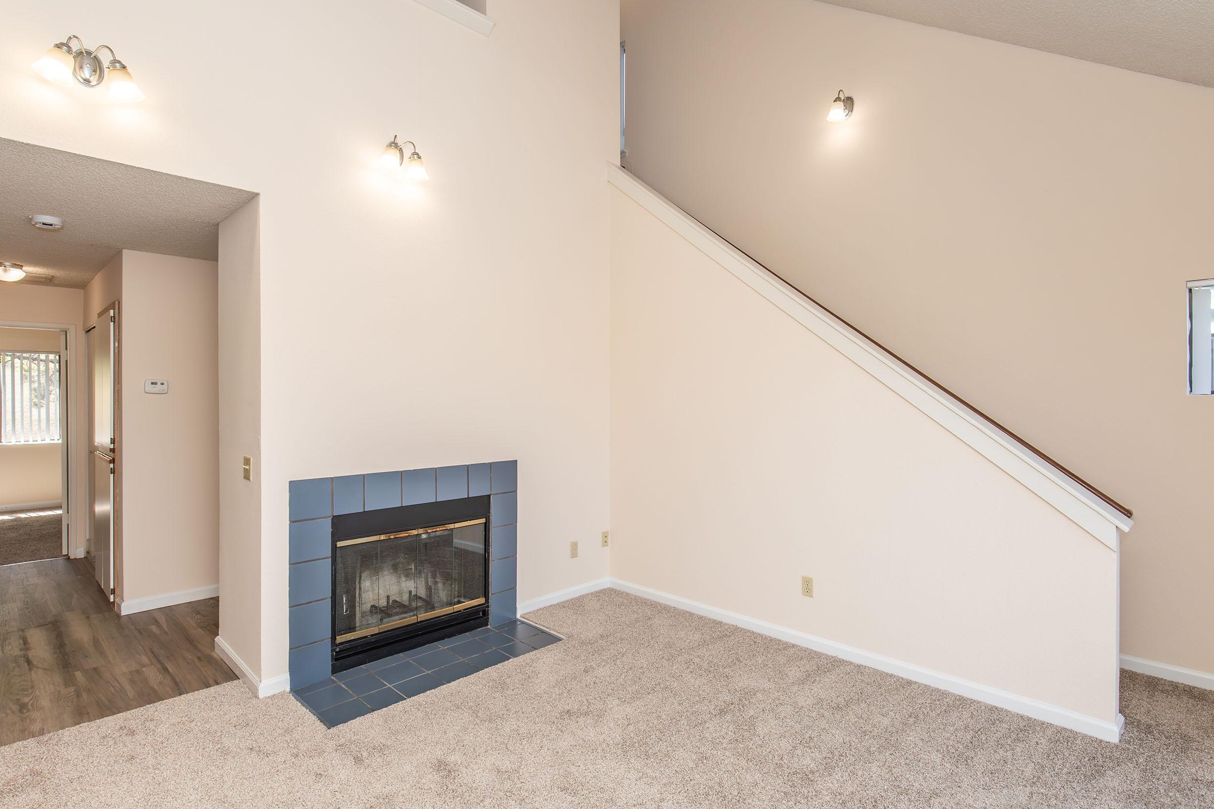 A spacious living area with light beige walls and carpet. There's a blue-tiled fireplace on one side, and a staircase leading to the upper level. Natural light is coming from a window in the room, and doorways are visible on the left. The overall atmosphere is bright and inviting.