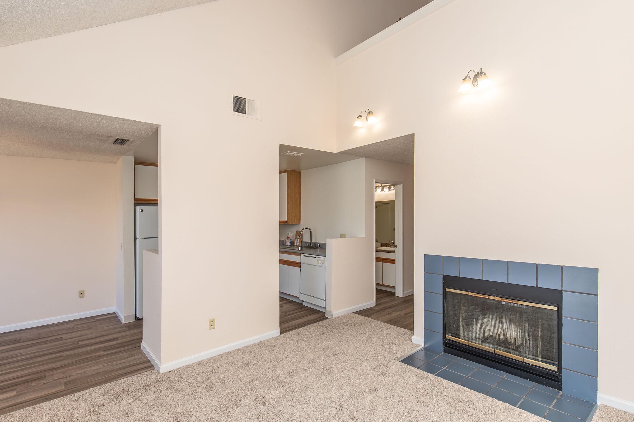 A spacious living area with beige walls, a blue-tiled fireplace, and carpeted flooring. To the left, an open kitchen area featuring wooden cabinets and white appliances is visible. Natural light enters through the ceiling height, making the room feel bright and airy.