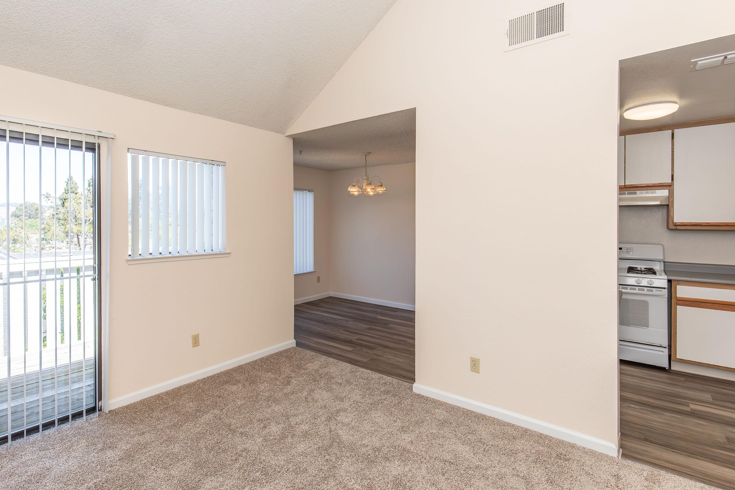 Spacious living area with carpeted flooring, featuring large windows with blinds that provide natural light. An open layout leads to a dining area with pendant lighting and an adjacent kitchen with white cabinets and appliances. Soft beige walls enhance the inviting atmosphere.
