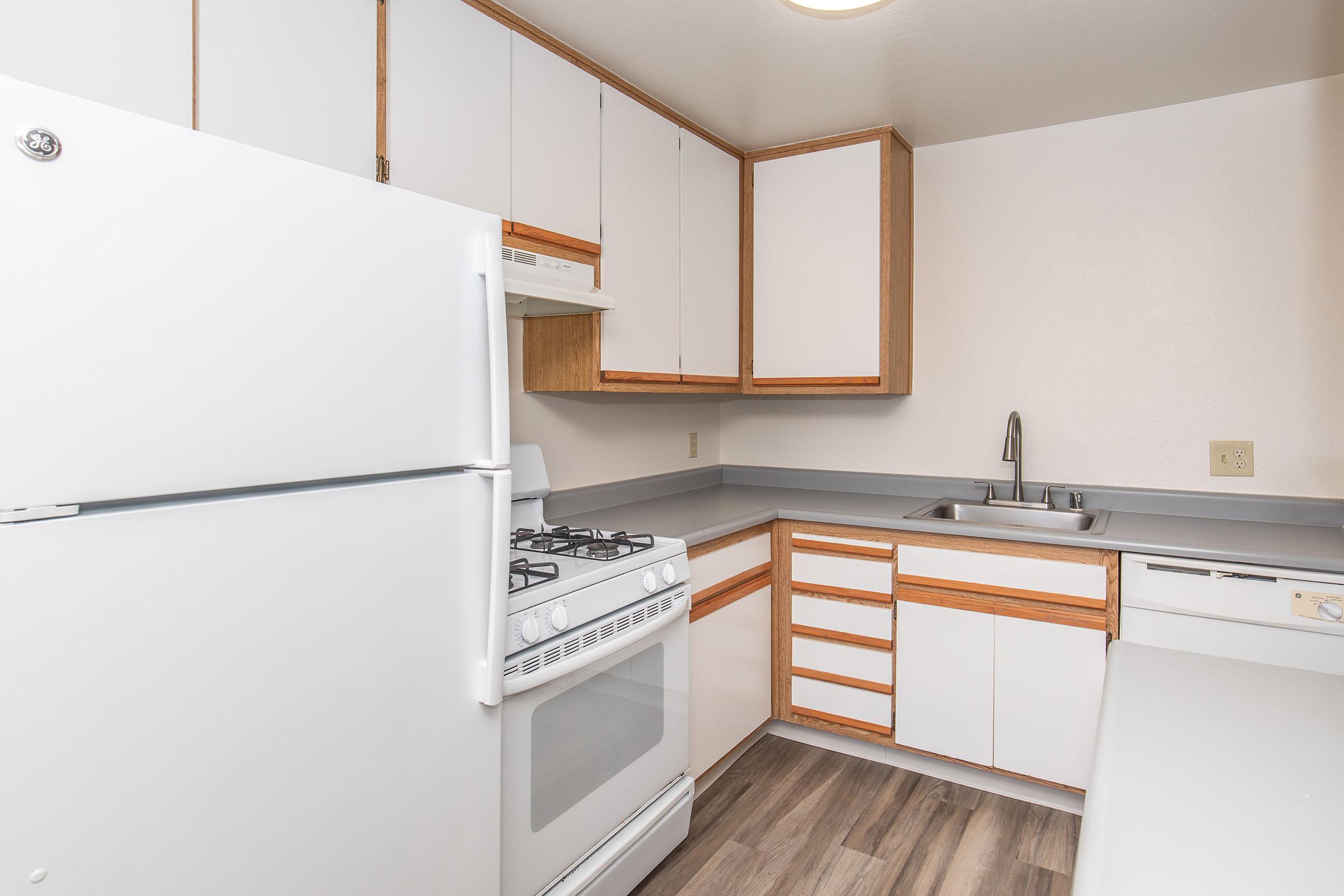 A clean, modern kitchen featuring white cabinets, a gas stove, and a large refrigerator. The countertop is gray, with a stainless steel sink and wooden accents. The flooring is a light wood laminate, giving the space a bright and open feel. The kitchen is well-organized and uncluttered.