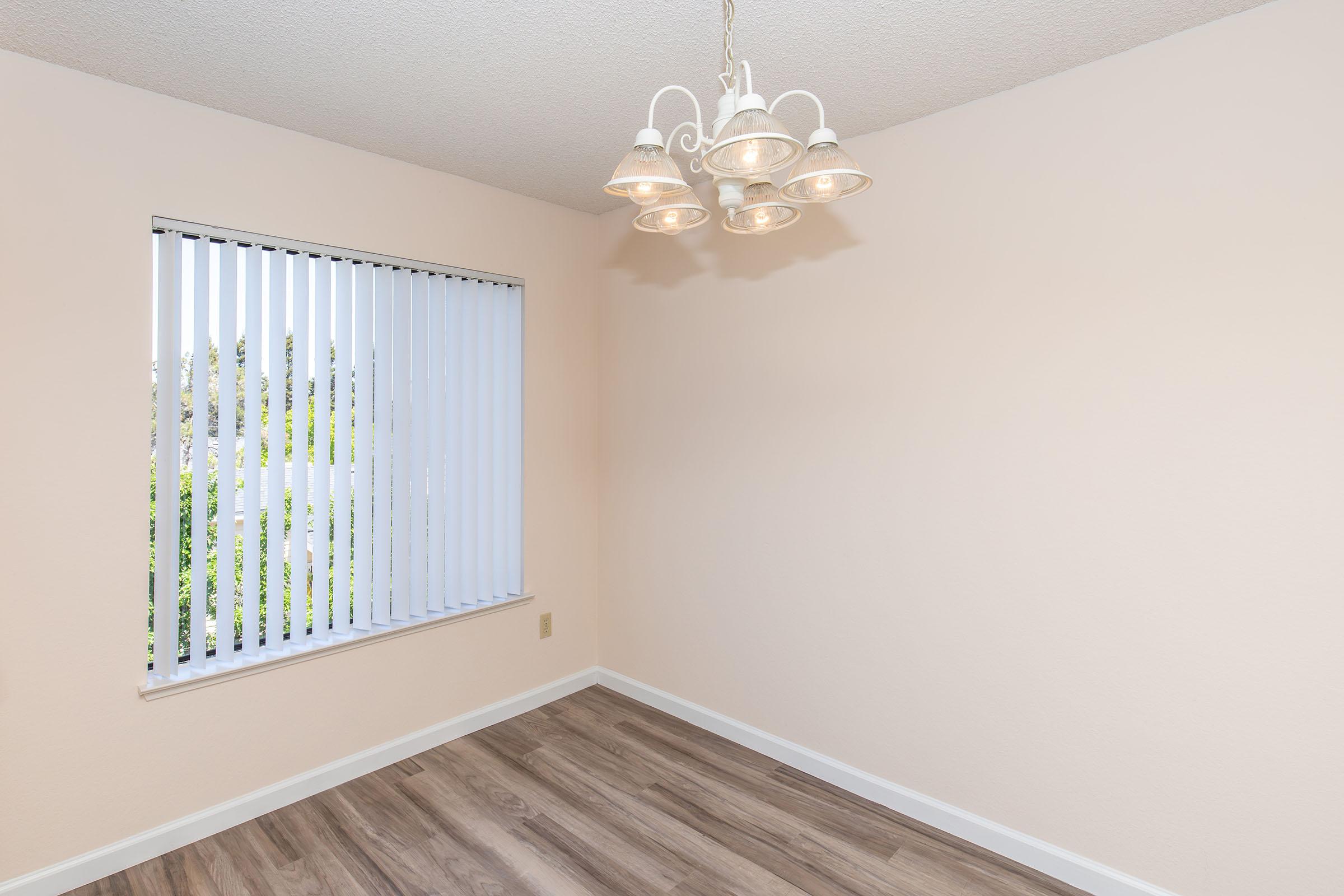 A softly lit empty room with light beige walls and a wood-look floor. A modern chandelier with multiple lights hangs from the ceiling. One window features vertical blinds, revealing greenery outside. The overall ambiance is calm and uncluttered, ideal for various uses.