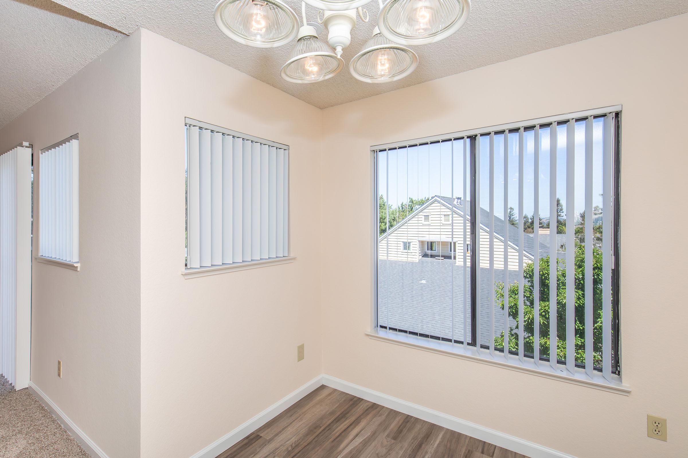 A well-lit corner of a room featuring beige walls, vertical blinds on two windows, and a modern light fixture with multiple bulbs. The floor is covered in wood-like laminate, and there is a view of greenery and neighboring buildings through the windows.