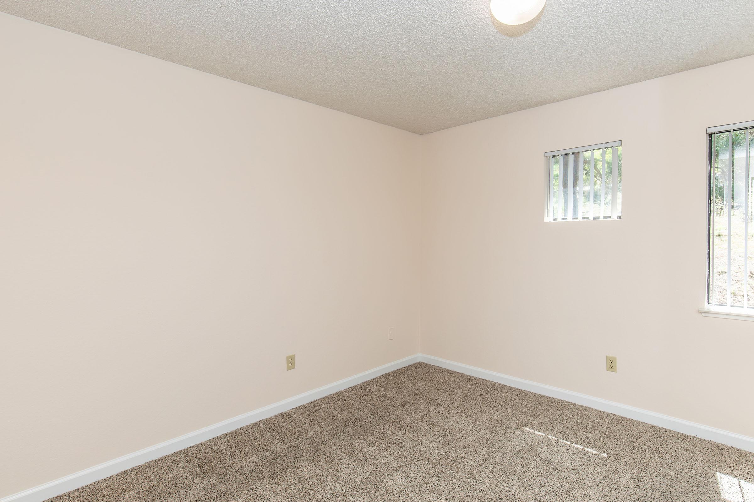 A vacant room featuring light beige walls and a tan carpet. The room has one small window with horizontal blinds, allowing natural light to enter. The ceiling is textured, and a light fixture is visible in the center. The space is empty and minimalistic, suitable for various uses.