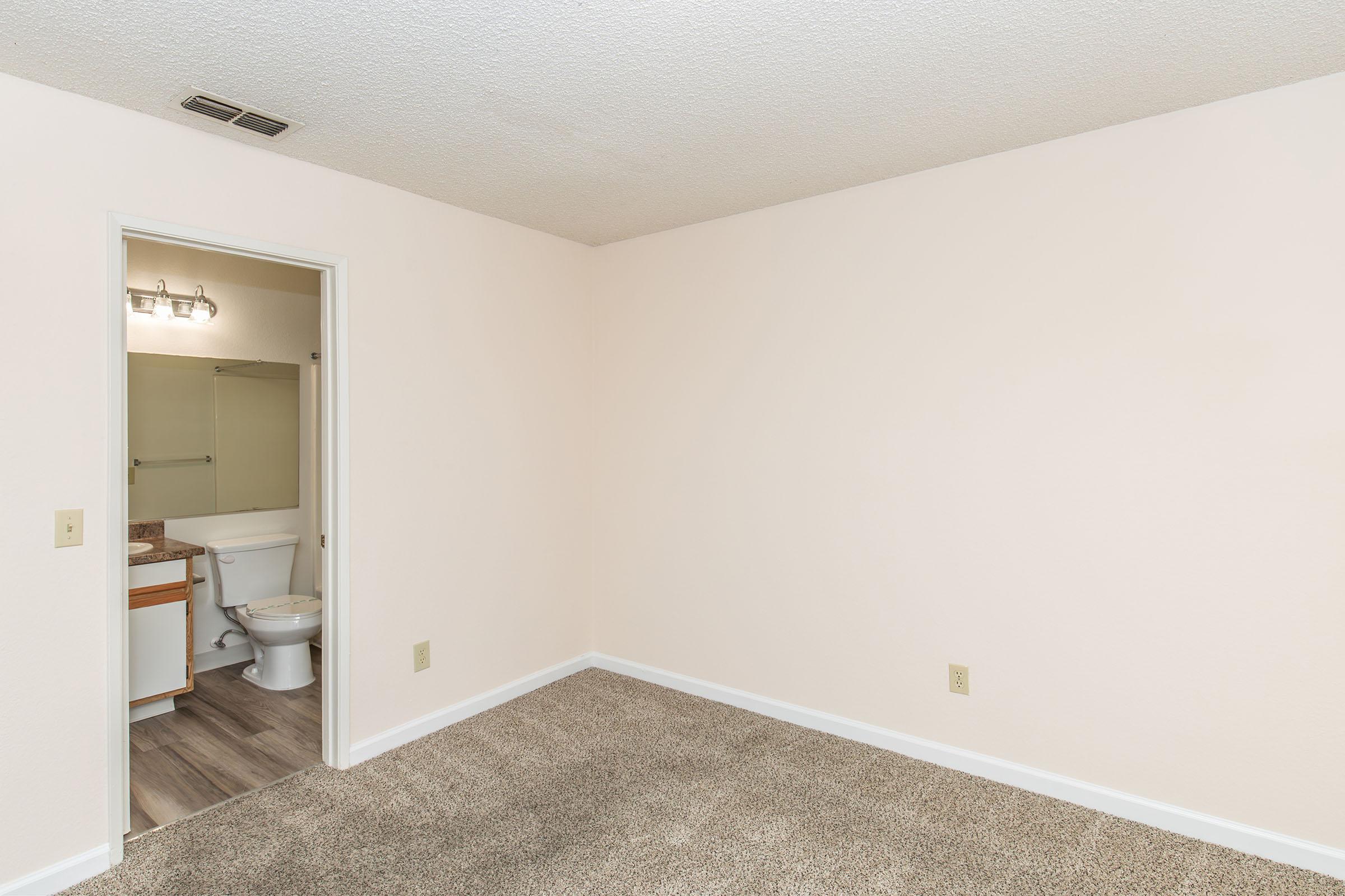 A sparsely furnished room with beige walls and carpet. A doorway leads to a bathroom featuring a toilet and sink. Natural light highlights the clean, minimalistic design, emphasizing the spaciousness of the area.