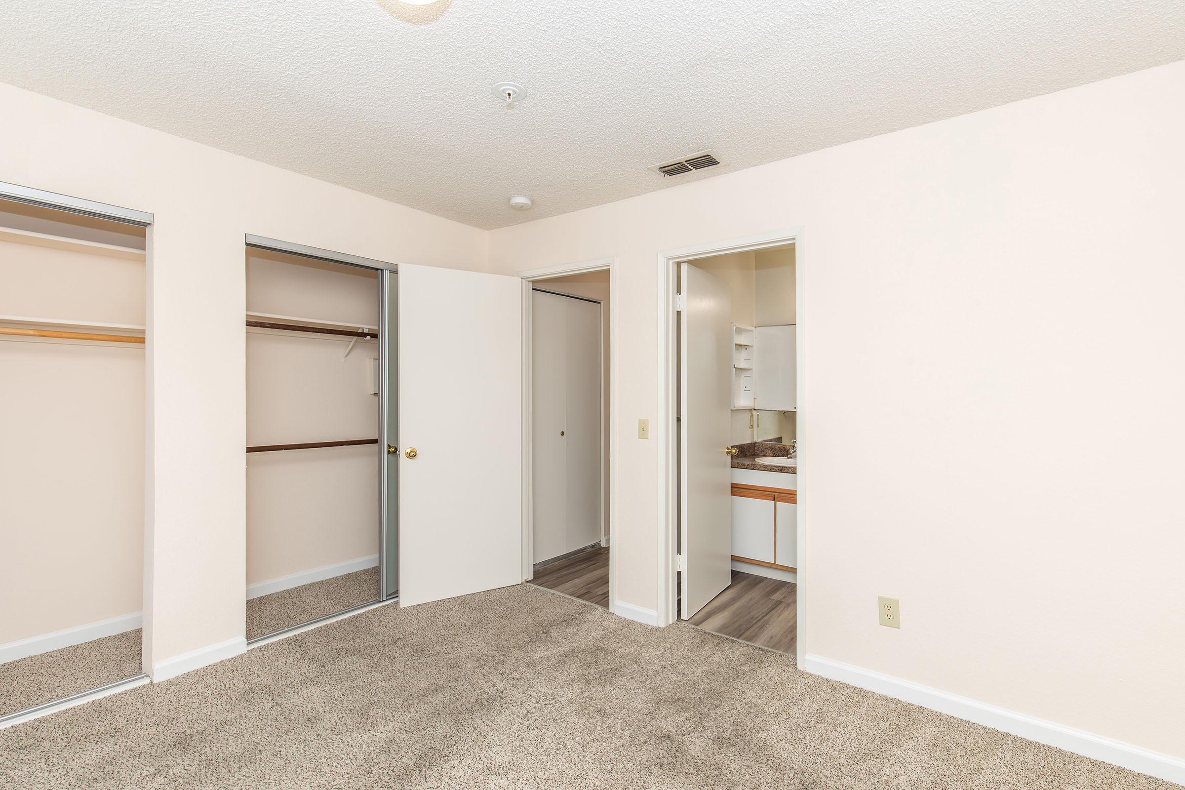 A view of a small, empty bedroom featuring light beige walls, carpeted flooring, and two open closet doors on the left. The room has a closed door leading to a bathroom area in the back, along with a light fixture on the ceiling. The space appears bright and clean, ready for occupancy.