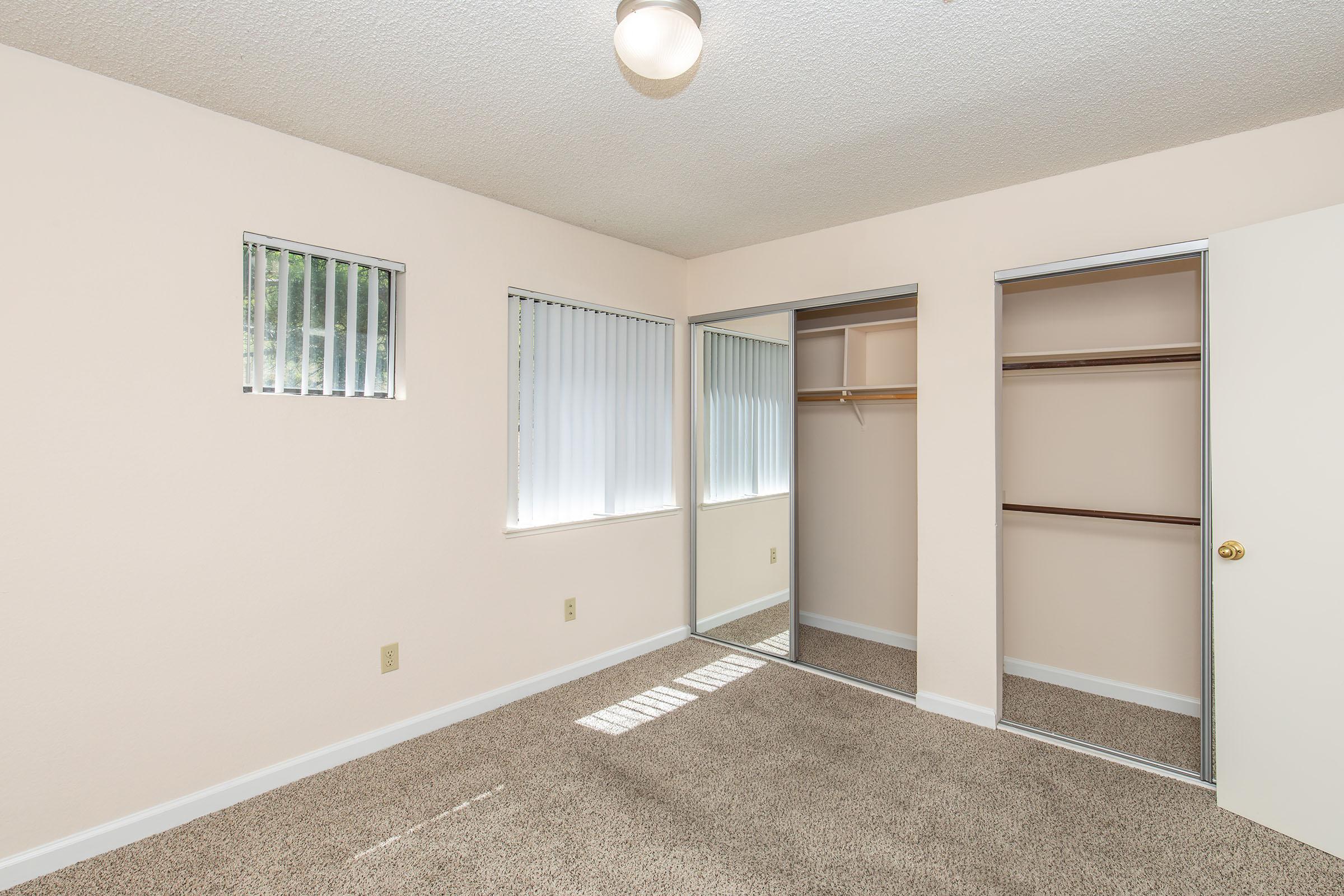 A well-lit bedroom featuring light beige walls and carpet. There are two small windows with blinds allowing natural light. A large mirrored closet with sliding doors and an open shelf is visible, alongside an interior door on the right. The overall atmosphere is clean and inviting.