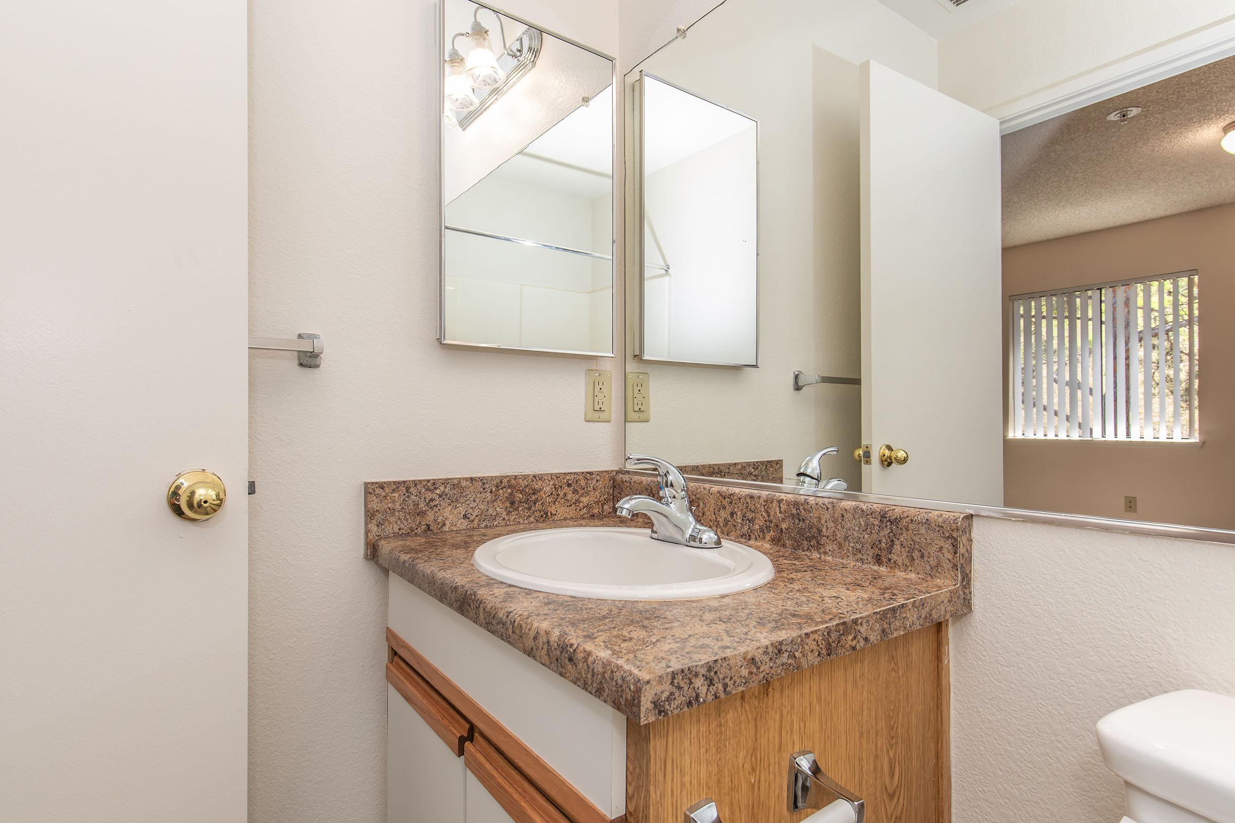 A clean bathroom with a light-colored walls and a granite countertop. There are two mirrors above the sink, and a faucet sits on the countertop. A towel rack is mounted on the wall, and there is a white toilet. A window with vertical blinds is visible in the background, providing natural light.