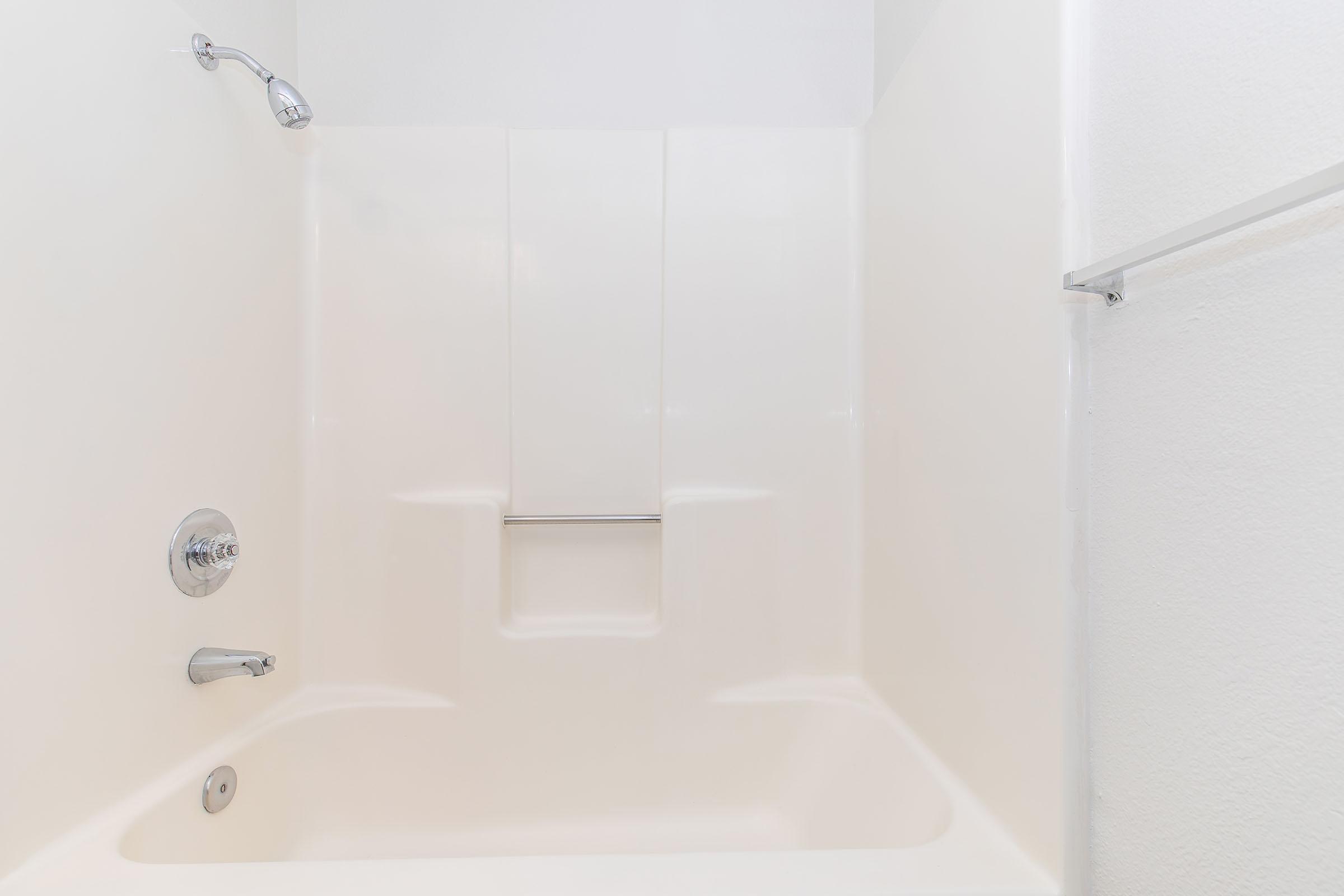 A clean, bright bathtub and shower area with white walls, featuring a chrome faucet and showerhead. The design includes a built-in seat and a grab bar for safety, creating a functional and accessible space. The overall look is modern and minimalistic.