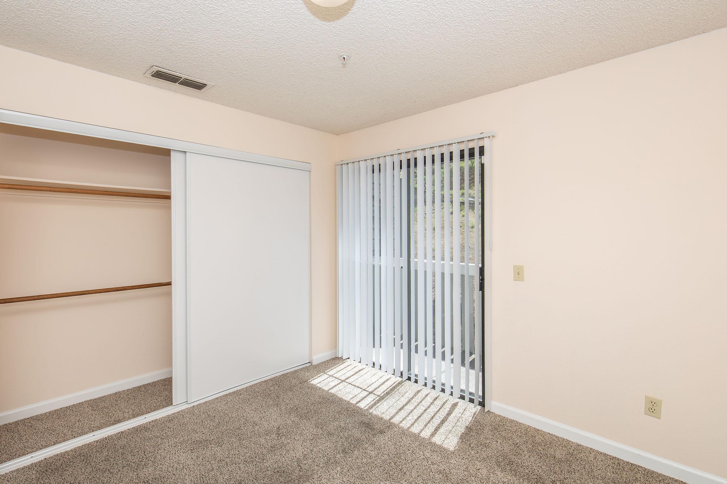 A bright, empty bedroom featuring light beige walls and carpet. There's a large sliding closet to the left, and a sliding glass door with vertical blinds leading outside. Sunlight streams in, creating a warm atmosphere. The room is sparsely furnished, emphasizing its spaciousness.