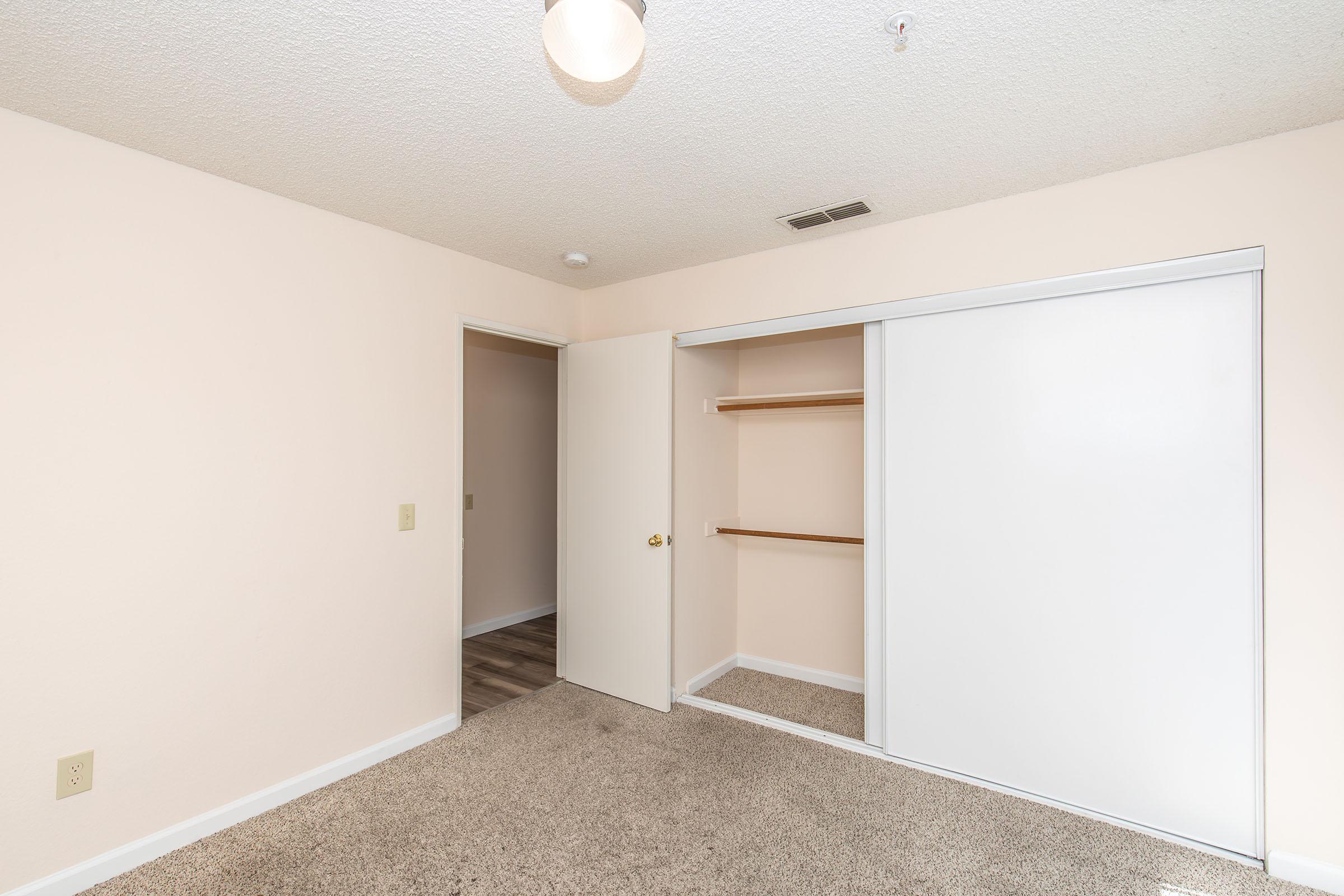 A lightly painted room with carpeted flooring. There's a closet with sliding white doors, featuring shelves. A door on the left leads to another room, and a ceiling light fixture is visible. The room appears clean and empty, suitable for various uses.