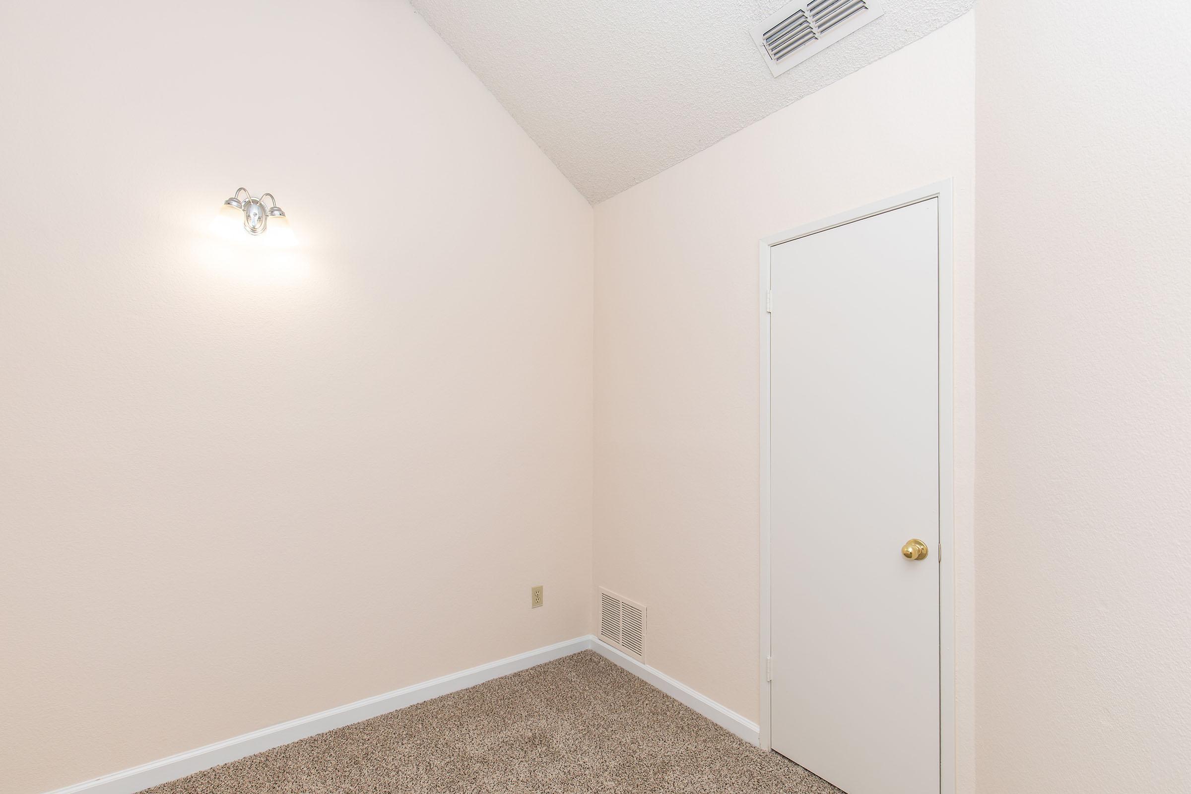 A small, empty room featuring a sloped ceiling, light beige walls, and carpeted flooring. There is a single light fixture on one wall and a closed white door on another wall. An air vent is visible near the floor.