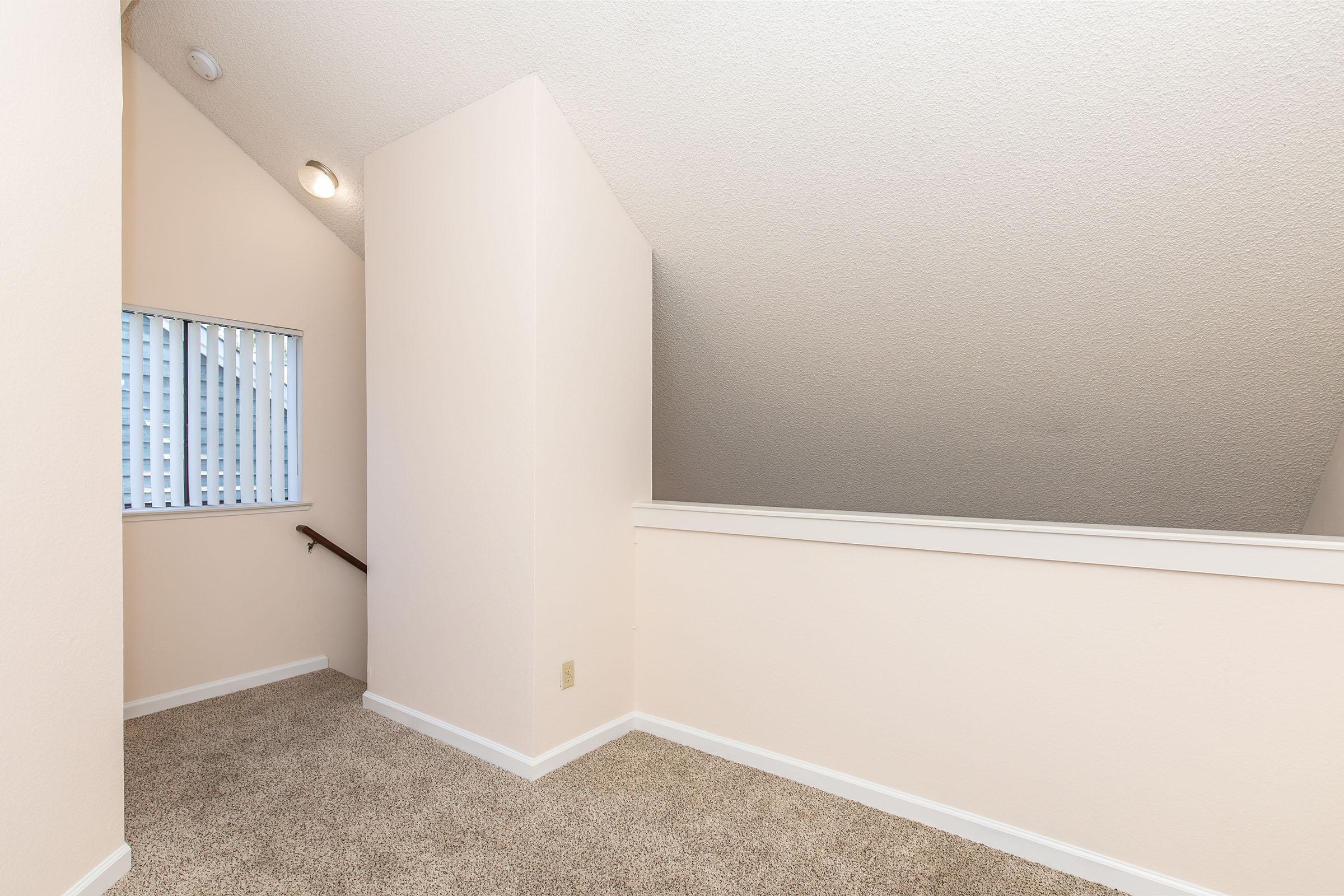A well-lit, carpeted area with light beige walls, featuring a sloped ceiling. There's a window with vertical blinds on the left side and a staircase railing leading out of the frame. The space appears clean and inviting, suitable for a cozy nook or sitting area.