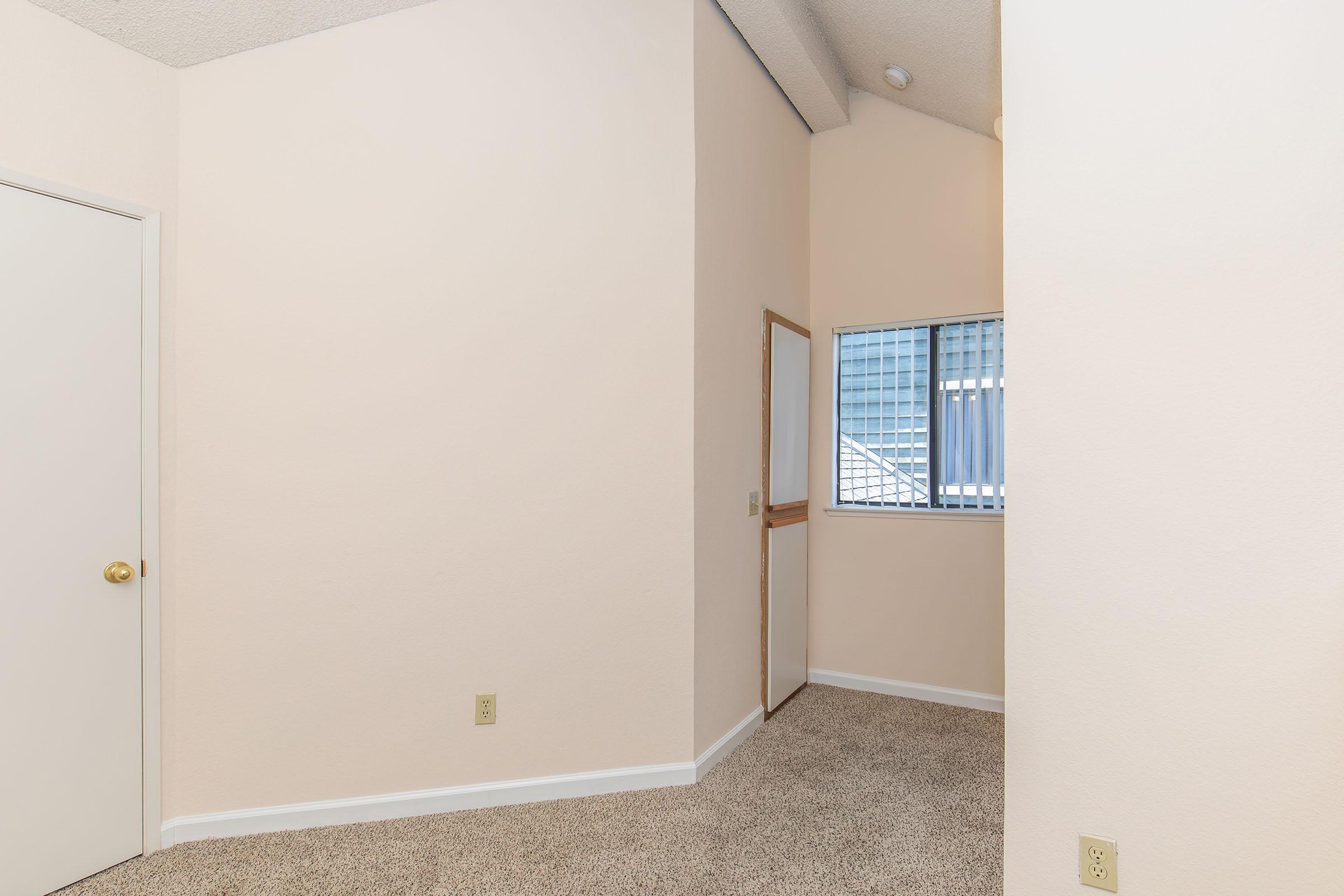 Interior of a room featuring light-colored walls and carpet. A door on the left leads to another area, while a small window is visible in the back, providing natural light. The space has a simple, clean design with a high ceiling.