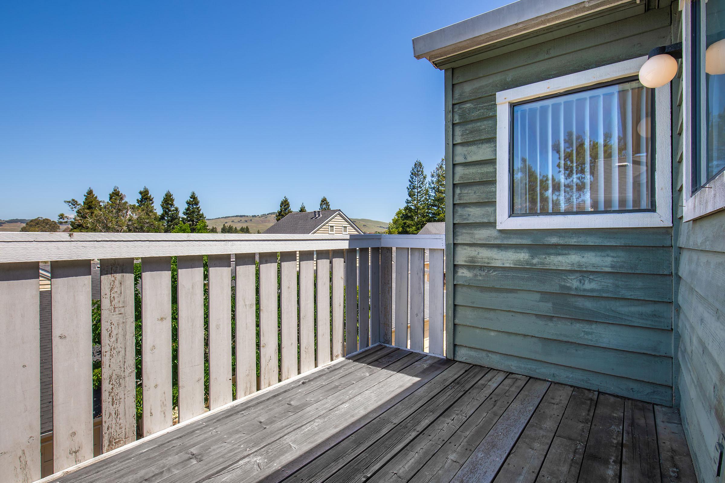 A wooden balcony with a gray wooden railing, overlooking a scenic view. The sky is clear and blue, and trees are visible in the background. A house can be seen in the distance, and sunlight casts a warm glow on the deck's surface. The balcony is attached to a building featuring green siding.